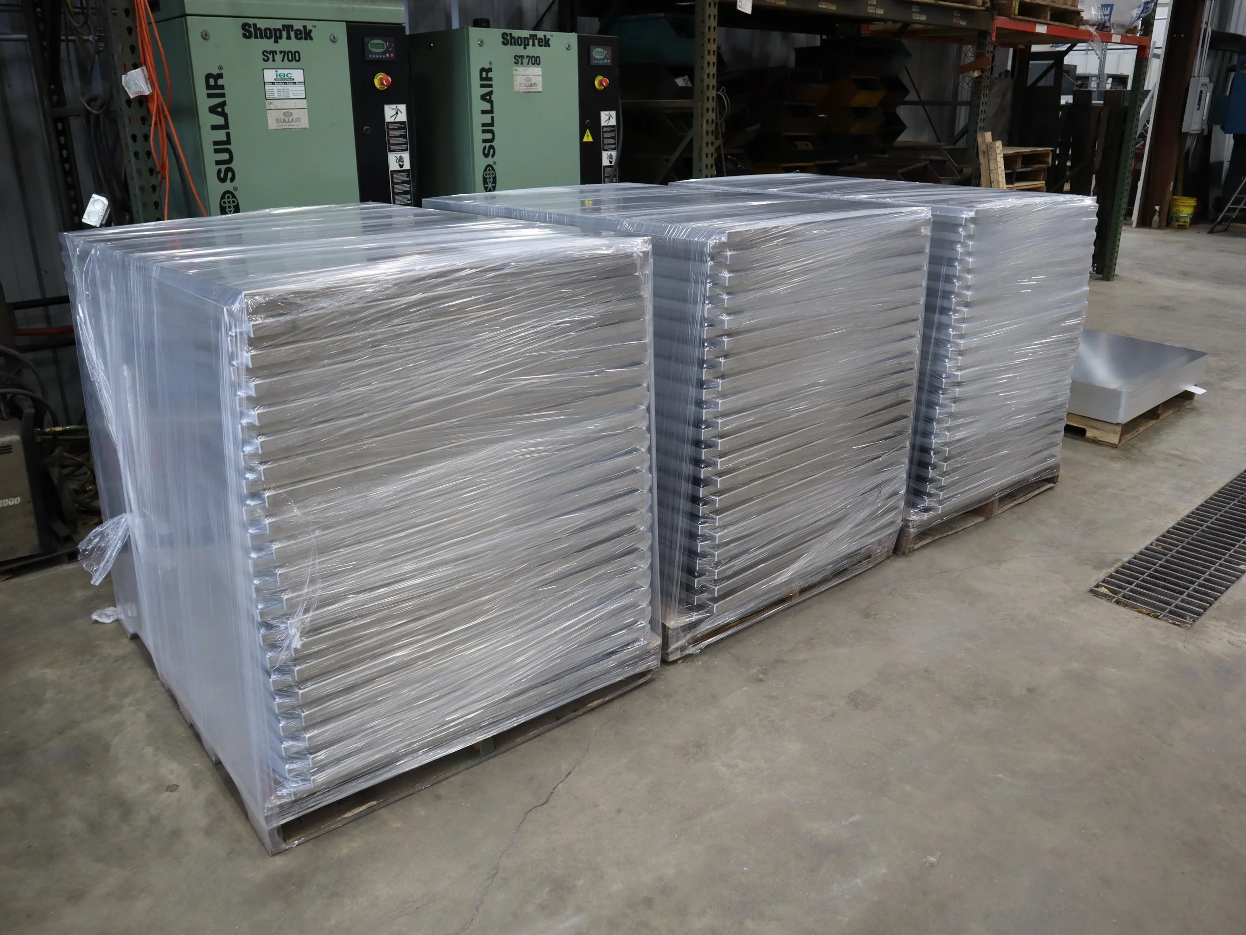 Three pallets of metal items wrapped in clear plastic in a warehouse. In the background, there are green air conditioning units and other industrial equipment.