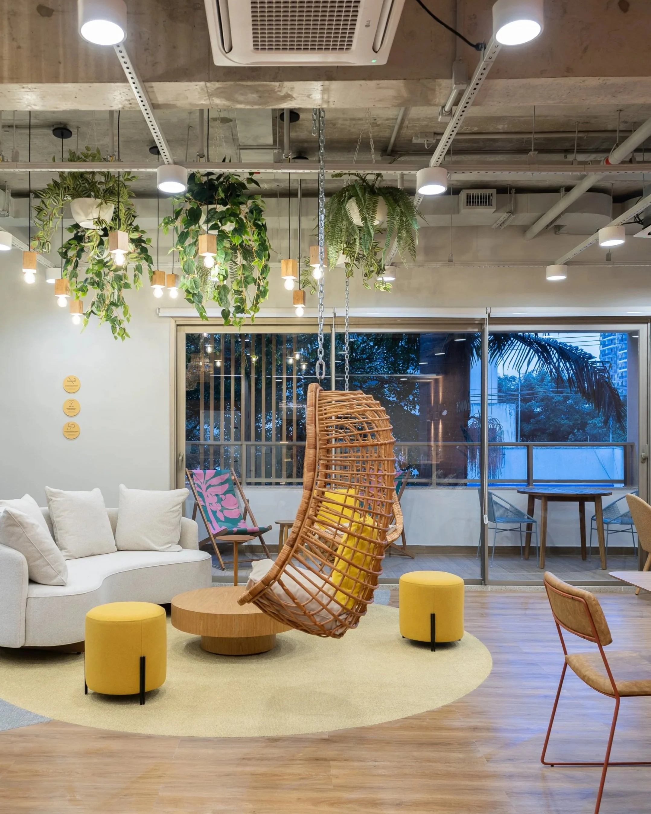 Sala de co-working moderna com poltronas de sofá branca, mesas pequenas amarelas, cadeiras de madeira, balanço de vime com almofadas amarelas, plantas suspensas e iluminação de teto.