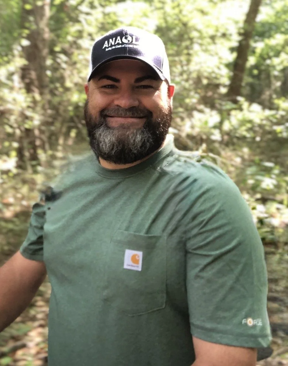 A smiling man with a beard wearing a baseball cap and green shirt outdoors in a forested area.