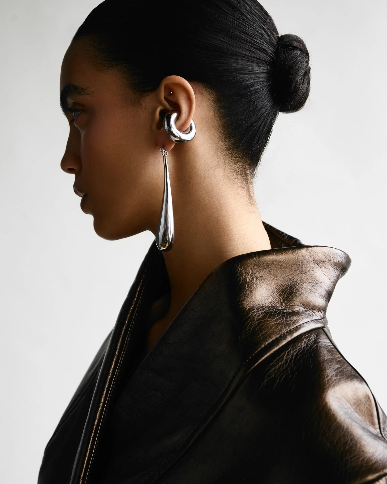 Profile of a woman with dark hair in a bun, wearing multiple silver earrings and a brown leather jacket.