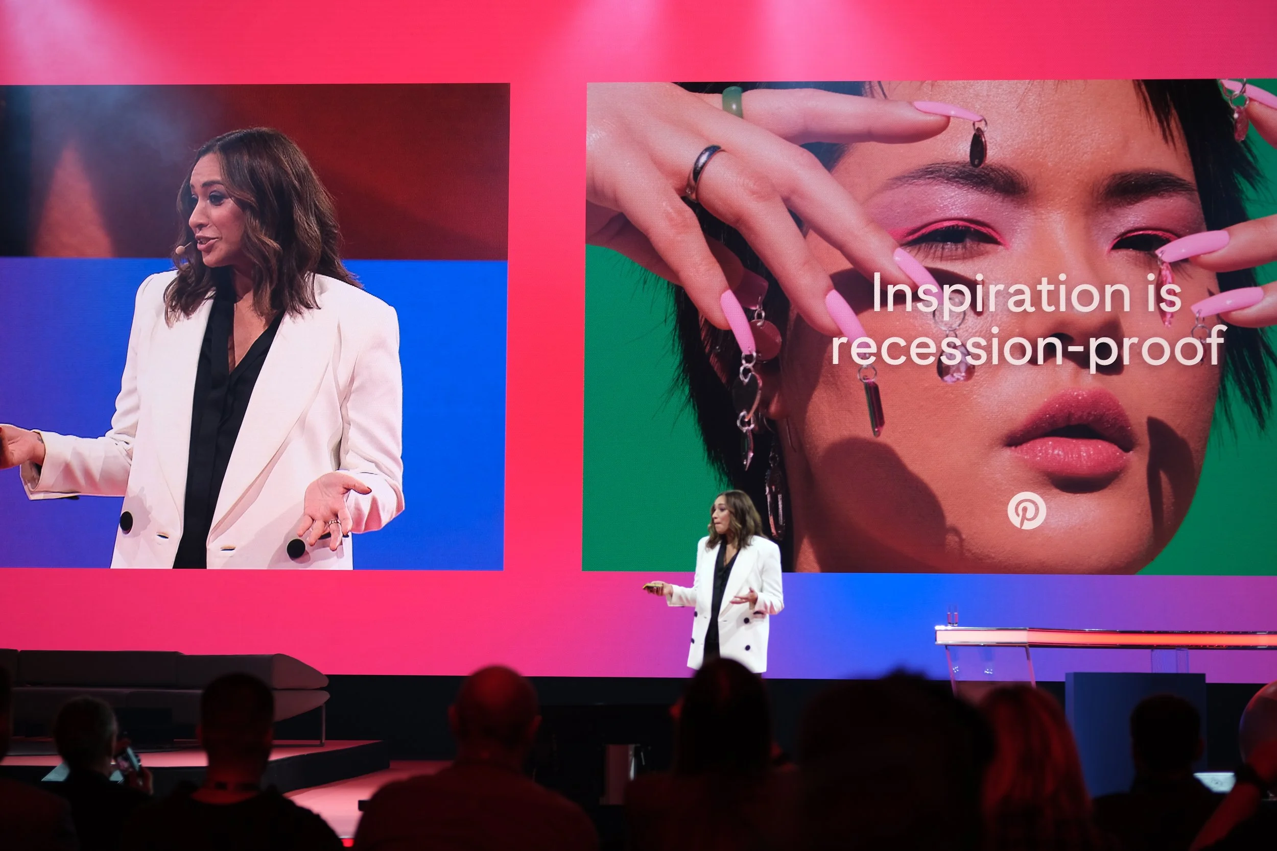 A woman is giving a presentation on stage with a large screen behind her displaying a close-up of a woman wearing makeup and jewelry. The slide includes the text "Inspiration is recession-proof." Audience members are visible in the foreground.