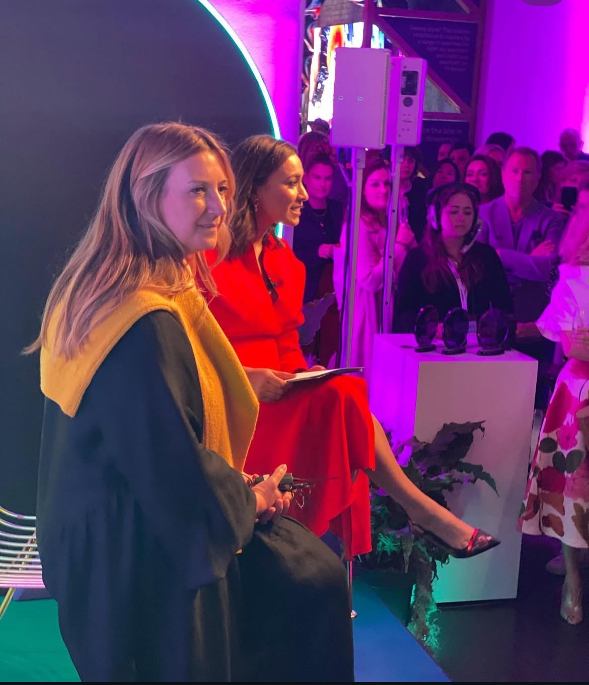 Women attending a presentation or panel discussion at an event with purple and pink lighting, and a group of people in the background.