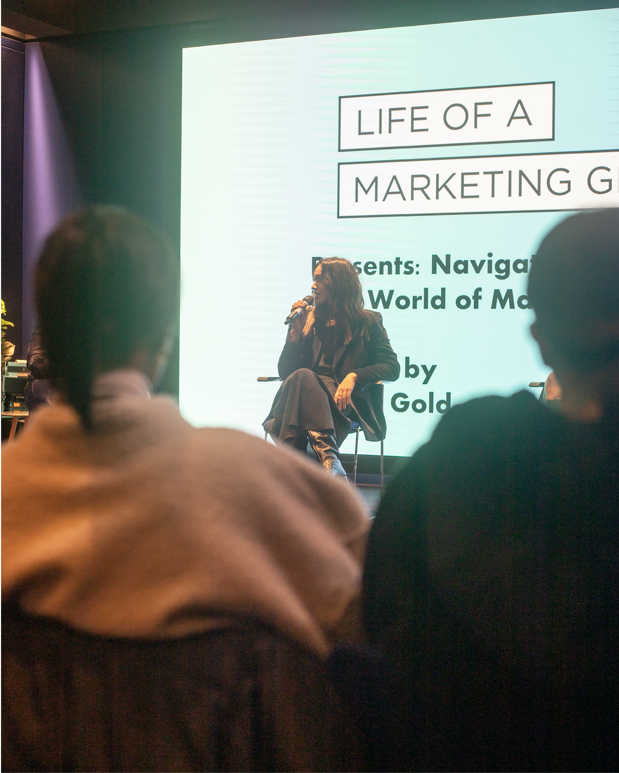 A woman speaking into a microphone at a conference stage, with a large screen behind her displaying the title 'Life of a Marketing Girl' and text about navigating the world of marketing.