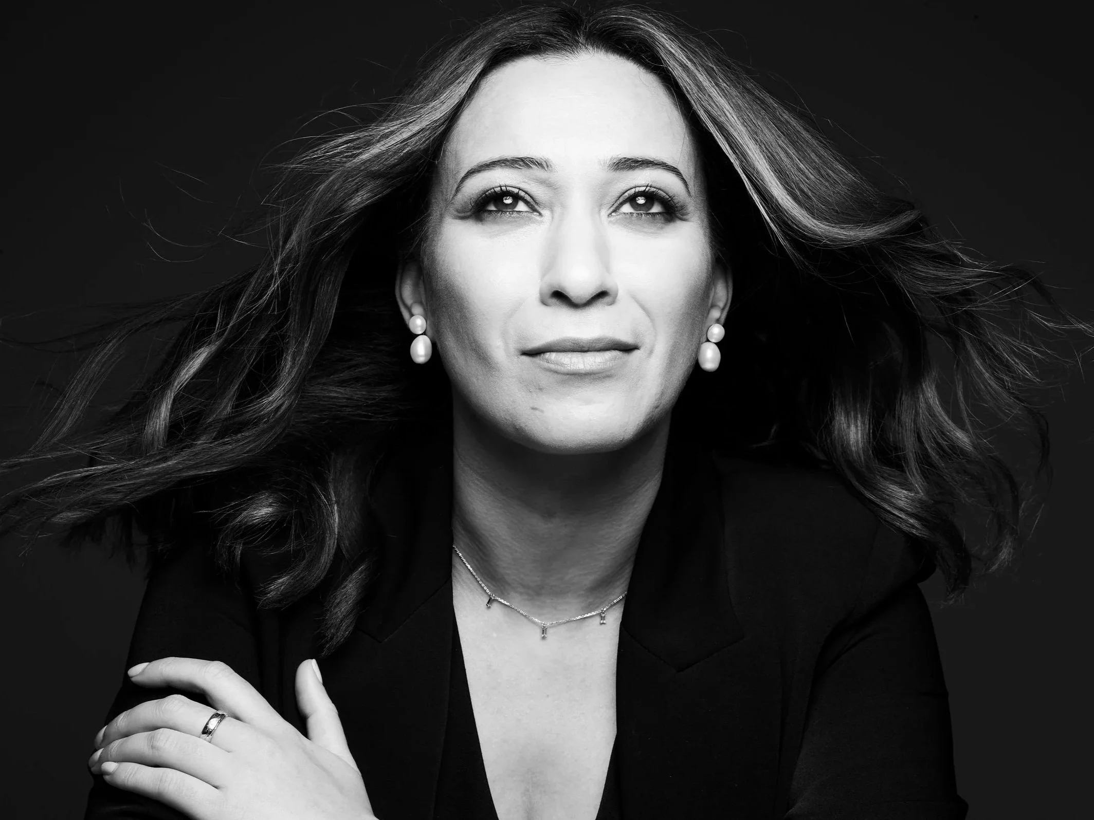 Black and white portrait of a woman with flowing hair, wearing earrings and a necklace, looking slightly upward with a confident expression.