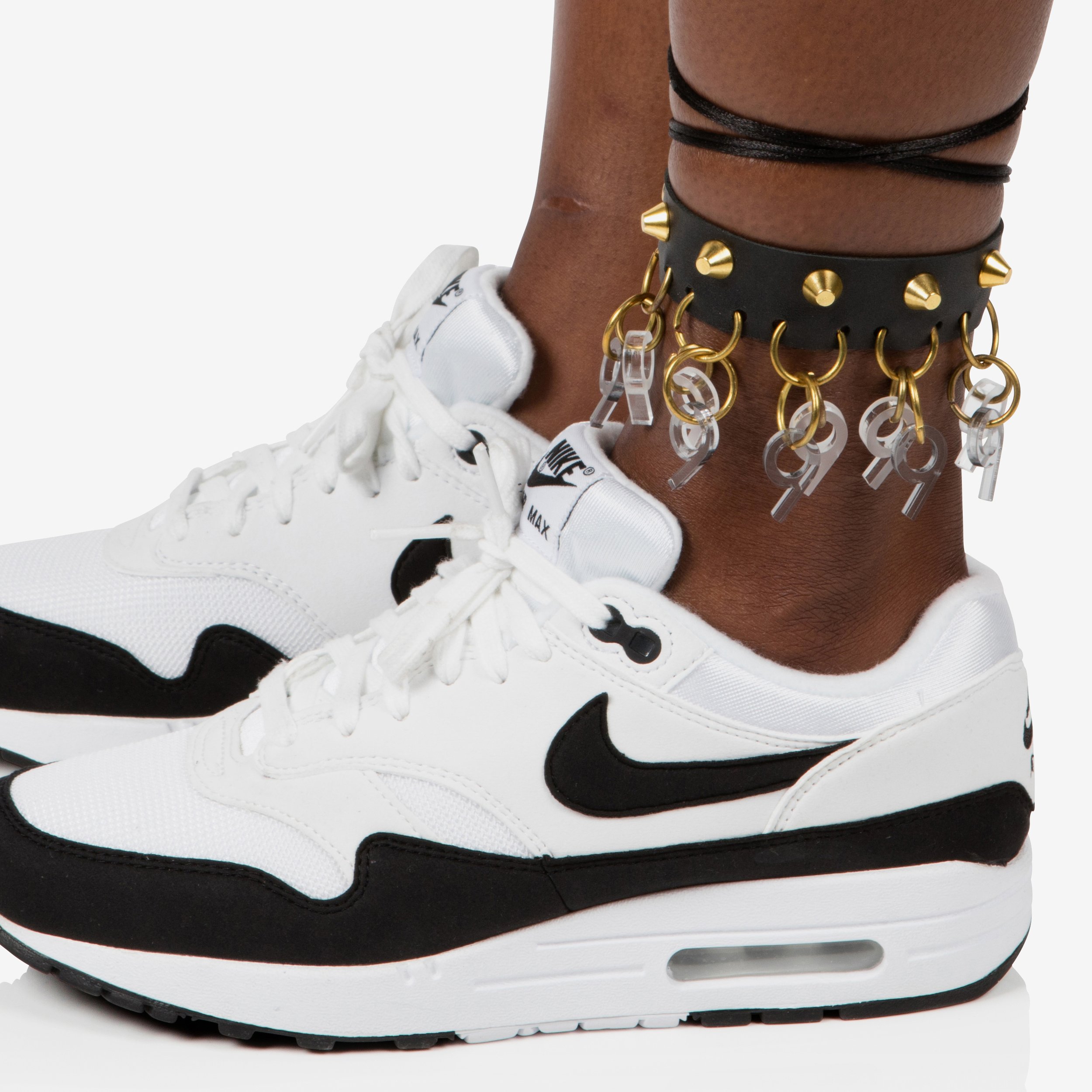 Close-up of a person's feet wearing white and black Nike sneakers with ankle jewelry, including a black leather strap with gold spikes and multiple gold and silver rings with charms.