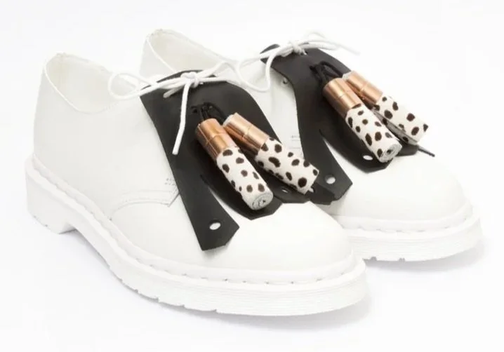Pair of unique white sneakers with black accents, decorated with leopard print laces and metallic copper-colored attachments on the laces.