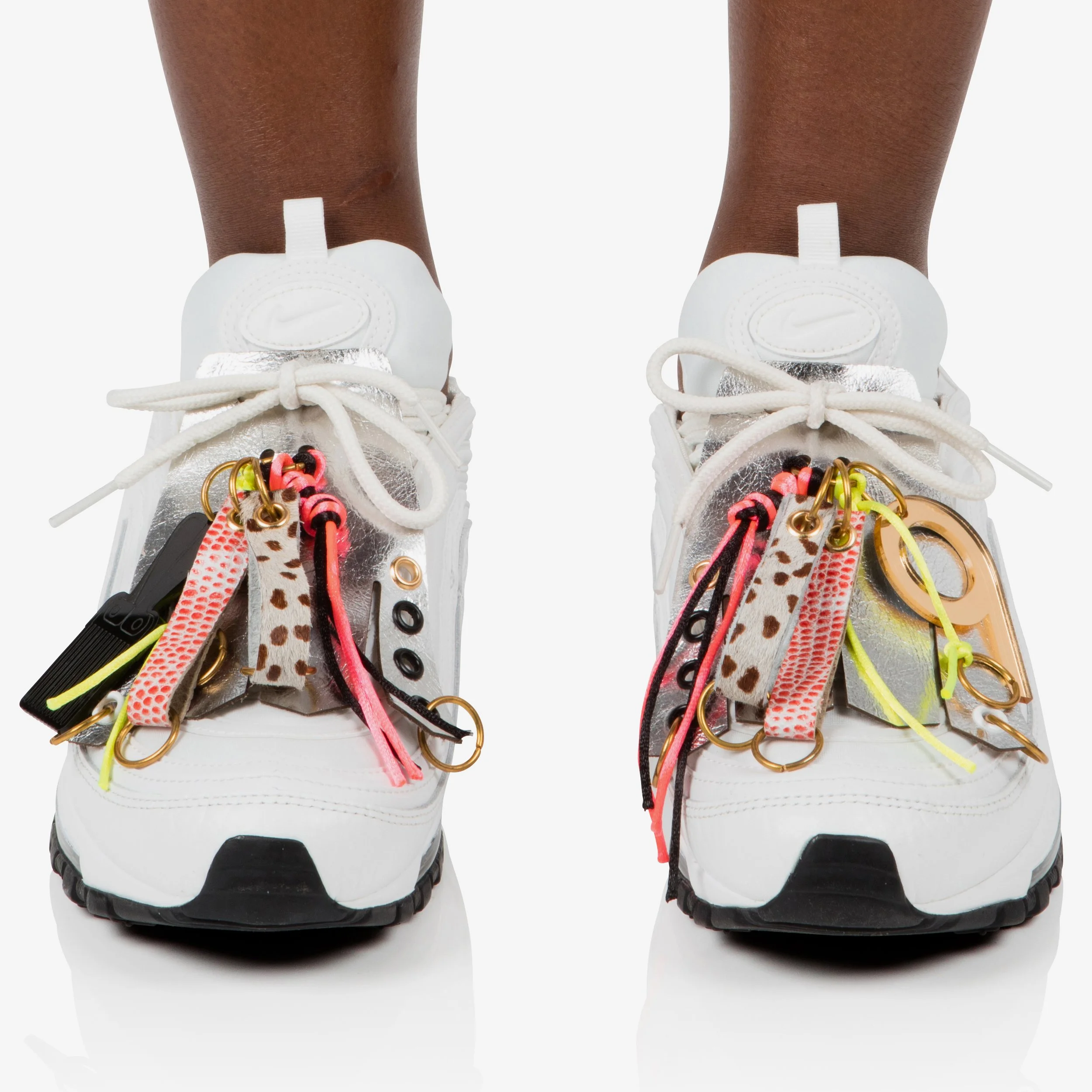 White Nike sneakers decorated with colorful and patterned shoe charms, including animal print, neon pink and yellow ribbons, and metallic accents.
