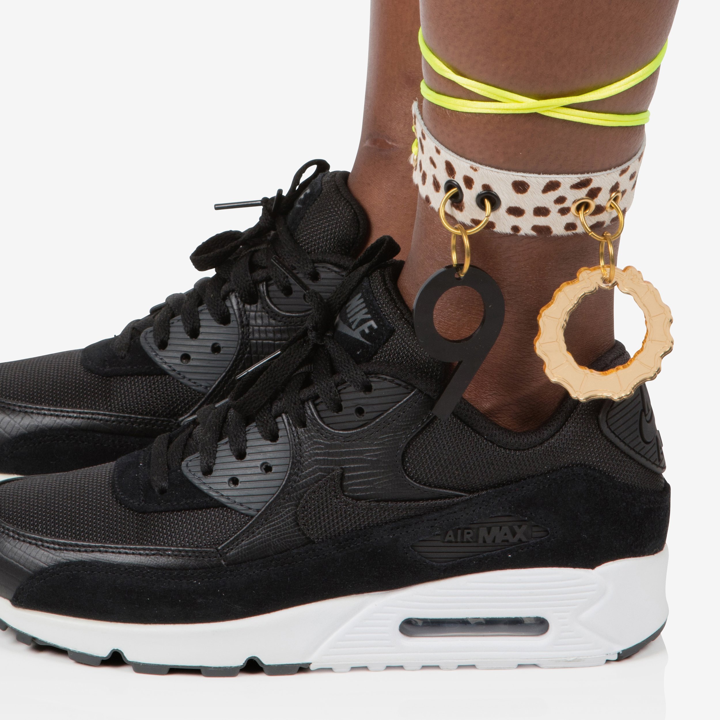 Close-up of a person's ankle wearing a black Nike Air Max sneaker, with colorful anklets and charms, against a white background.