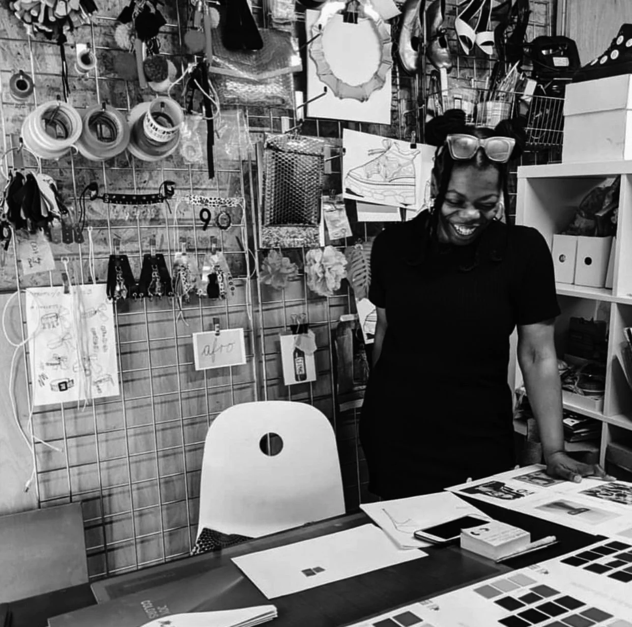 A woman with glasses on her head, smiling and looking down, standing in a creative workspace with craft and design supplies on the table and a wall filled with hanging jewelry, sketches, and decorative items.