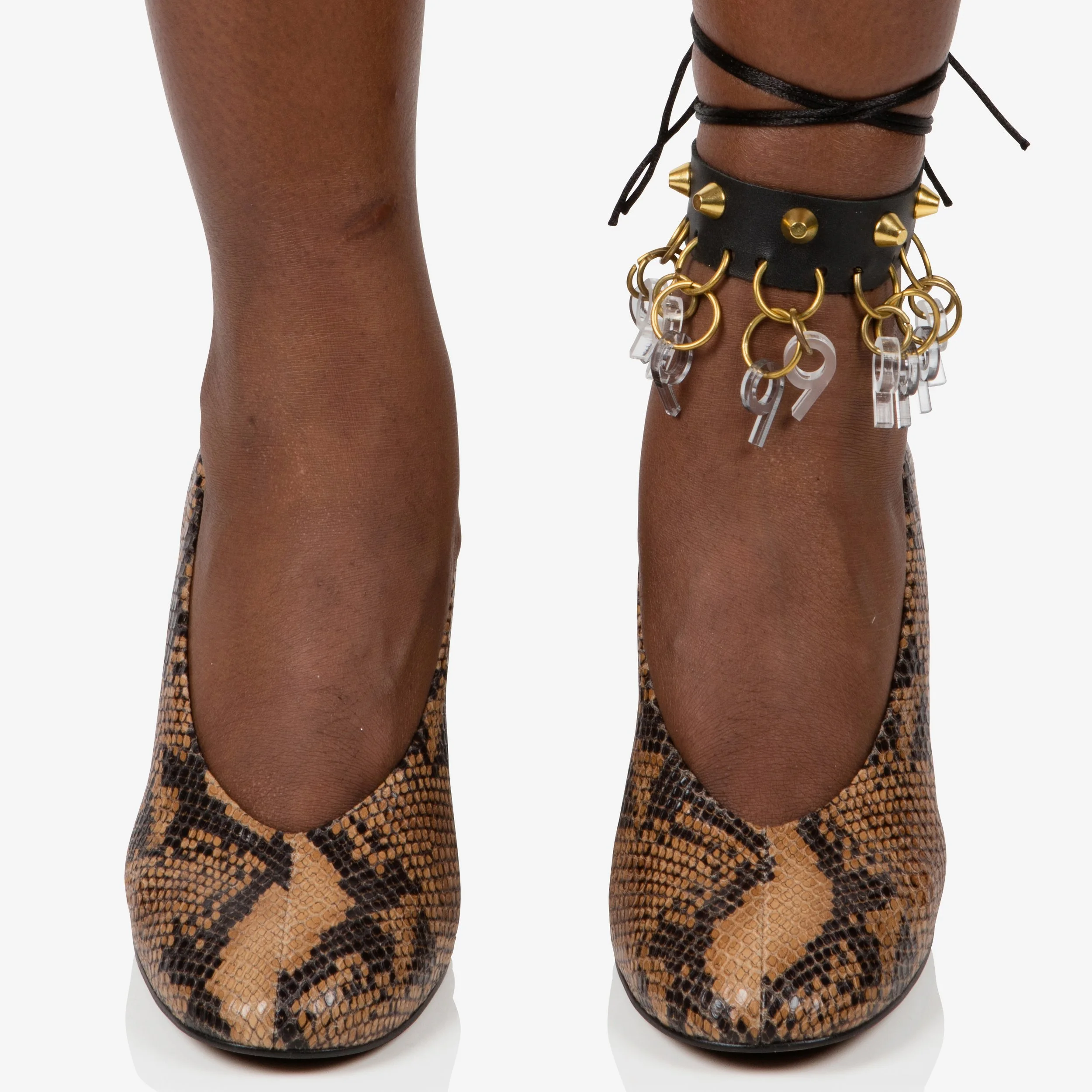 Close-up of a pair of high-heeled shoes with a snakeskin pattern, worn by a person with dark skin, featuring an ankle accessory with black leather, gold spikes, and multiple small key charms.