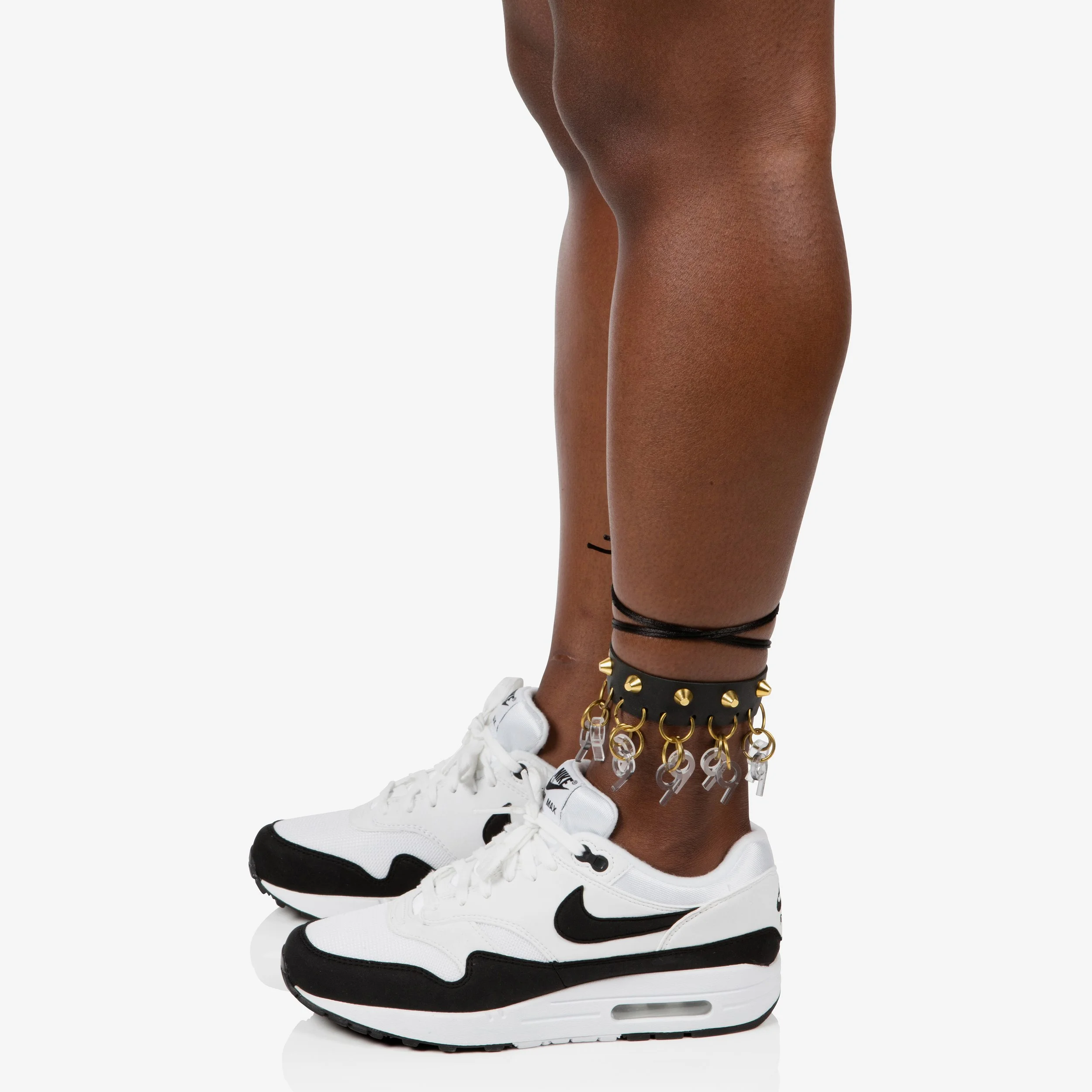 Person's legs with white Nike sneakers, black ankle jewelry, and anklets with various charms.