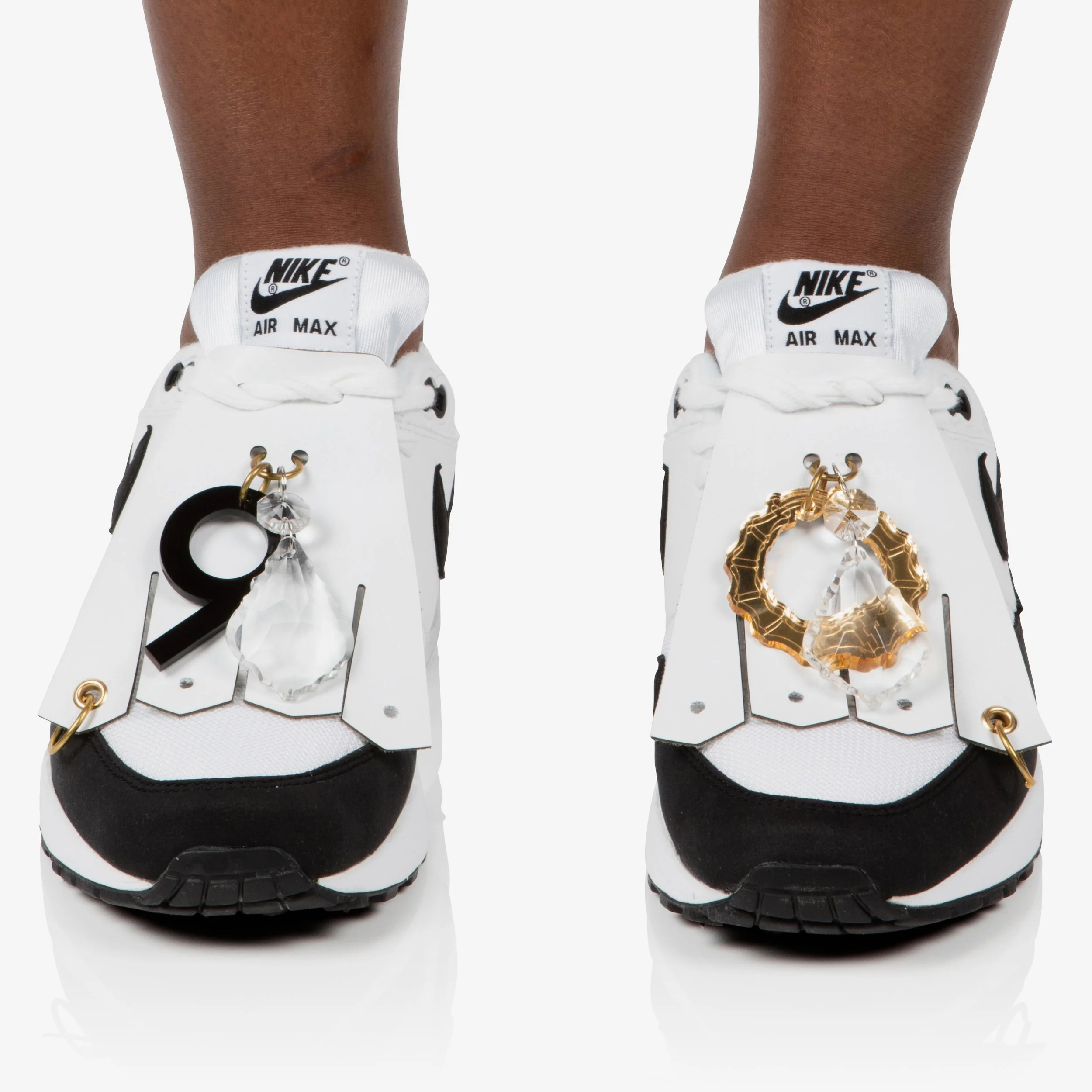 Pair of white Nike Air Max sneakers with decorative metal and crystal charms hanging from the laces, one black and one gold, on each shoe.