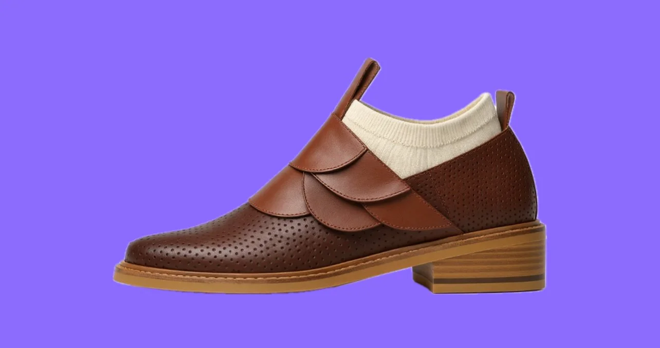 Brown leather shoe with decorative scalloped overlays, a low block heel, and a beige sock against a purple background.