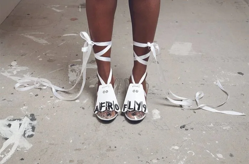 Close-up of a person's feet wearing white high-heeled shoes with the words 'Aero Fly' written on the toes, and white ribbon laces wrapped around the ankles, standing on a gray, painted, and slightly stained floor.