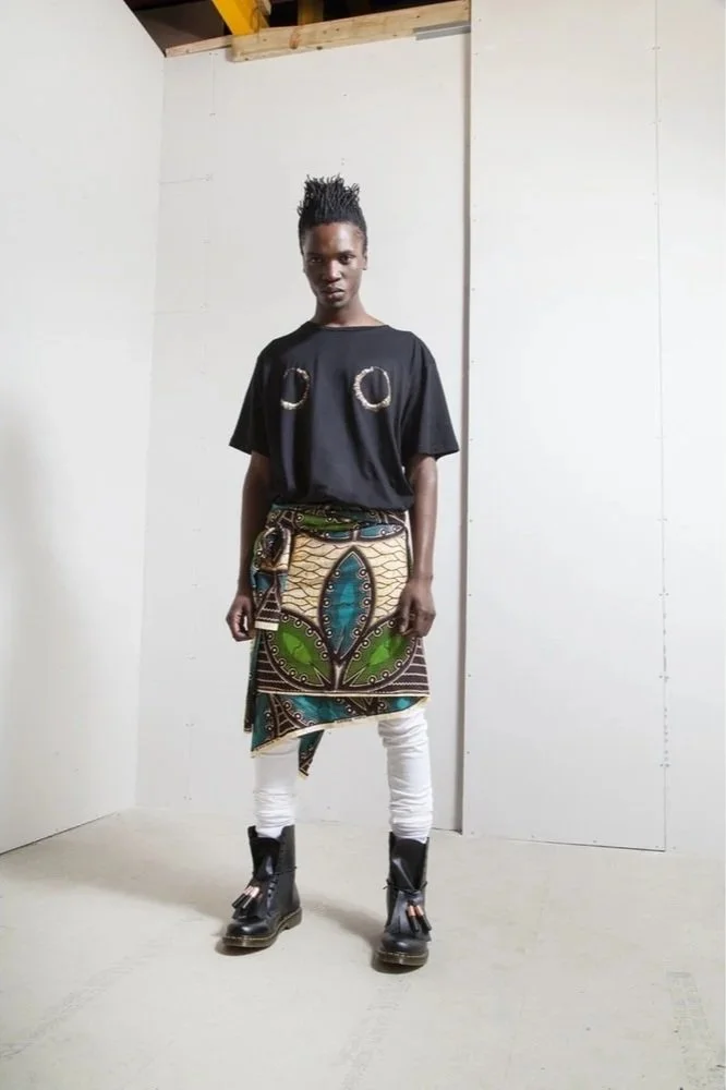 Person standing in a plain room, wearing a black t-shirt with metallic designs on the chest, a colorful patterned skirt, white leggings, and black boots.