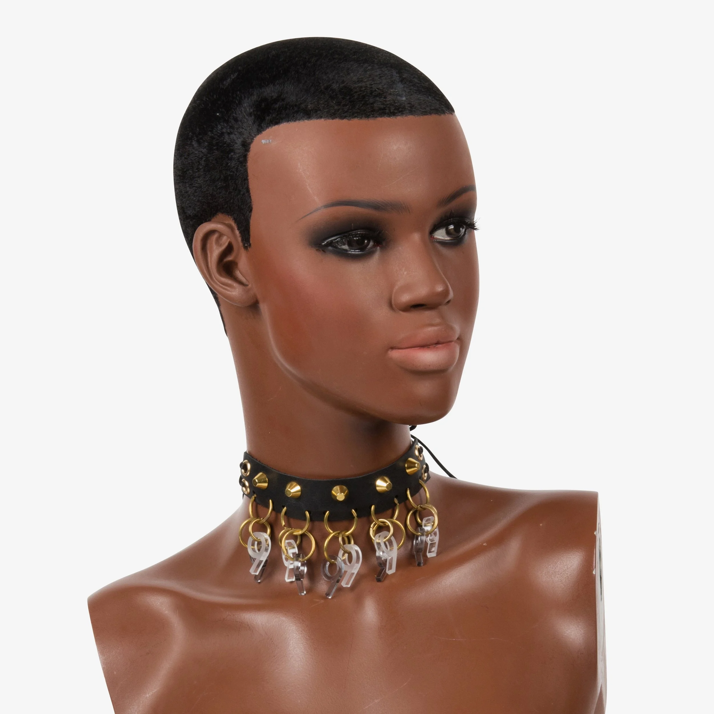 A mannequin with dark skin, a clean haircut, wearing dark eye makeup, an edgy black choker with gold studs, silver and gold key charms, and a blank white background.
