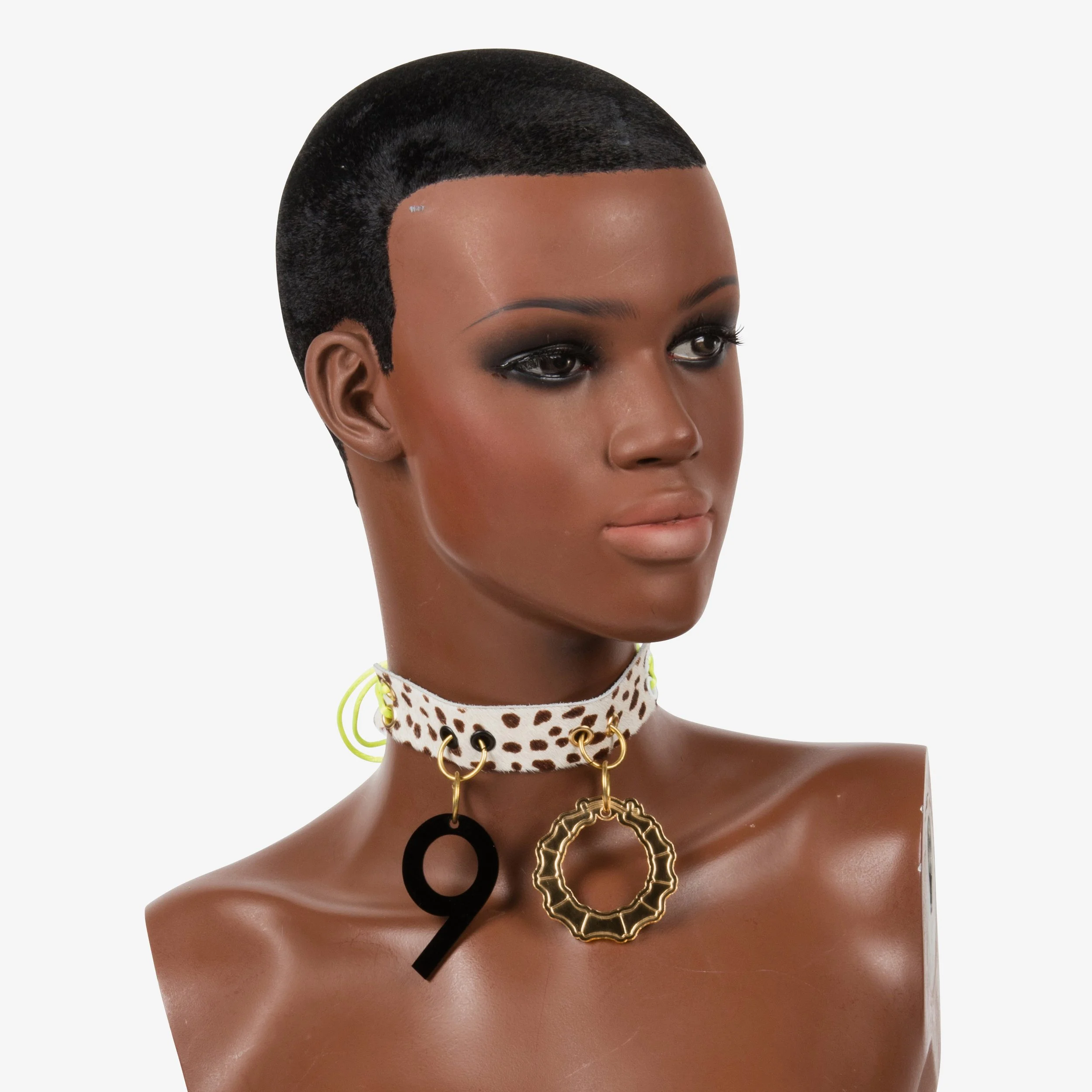 A mannequin with dark skin, short black hair, and wearing statement jewelry, including a chunky choker with a large black '9' and a gold circular pendant, against a plain white background.