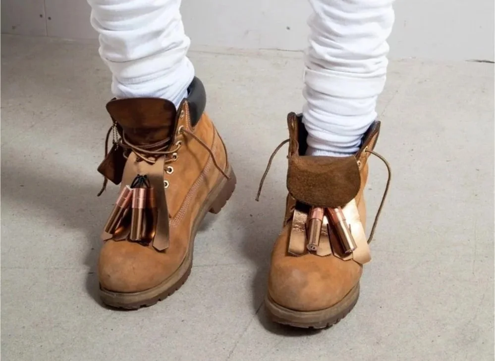 Pair of tan work boots with copper-colored nuts and bolts, worn with white pants, on a concrete floor.