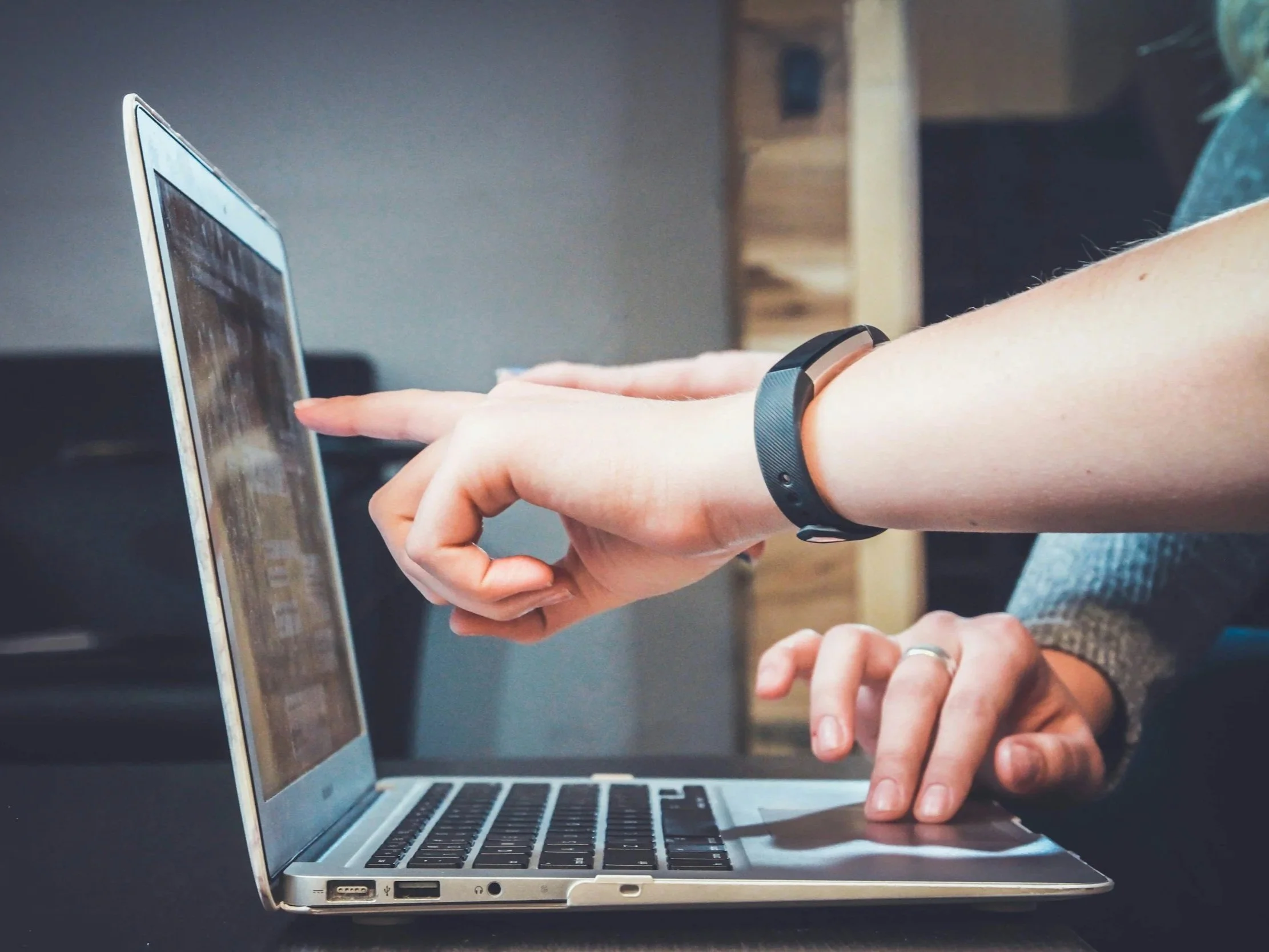 Person using a laptop with a smartwatch on their wrist, pointing at the laptop screen, in an indoor setting.