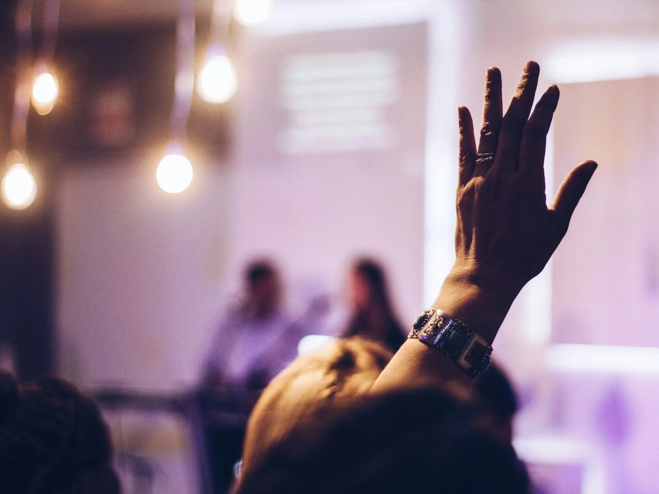 Close-up of a person raising their hand with an ornate bracelet, blurred background with two people, dim lighting and hanging light bulbs, possibly at a meeting or religious service.