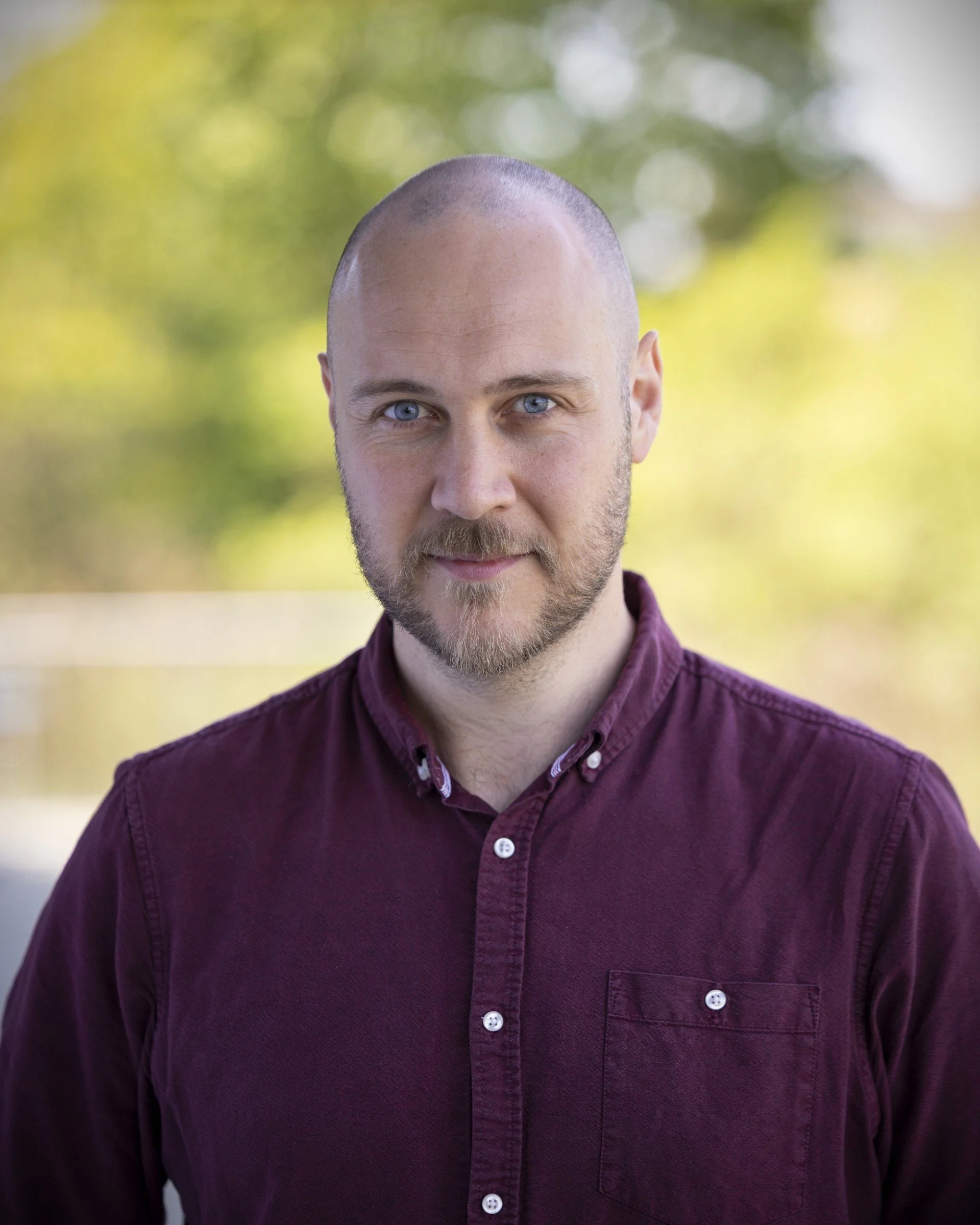 Portrait of a man with a beard and shaved head, wearing a maroon shirt, outdoors with blurred green foliage in the background.
