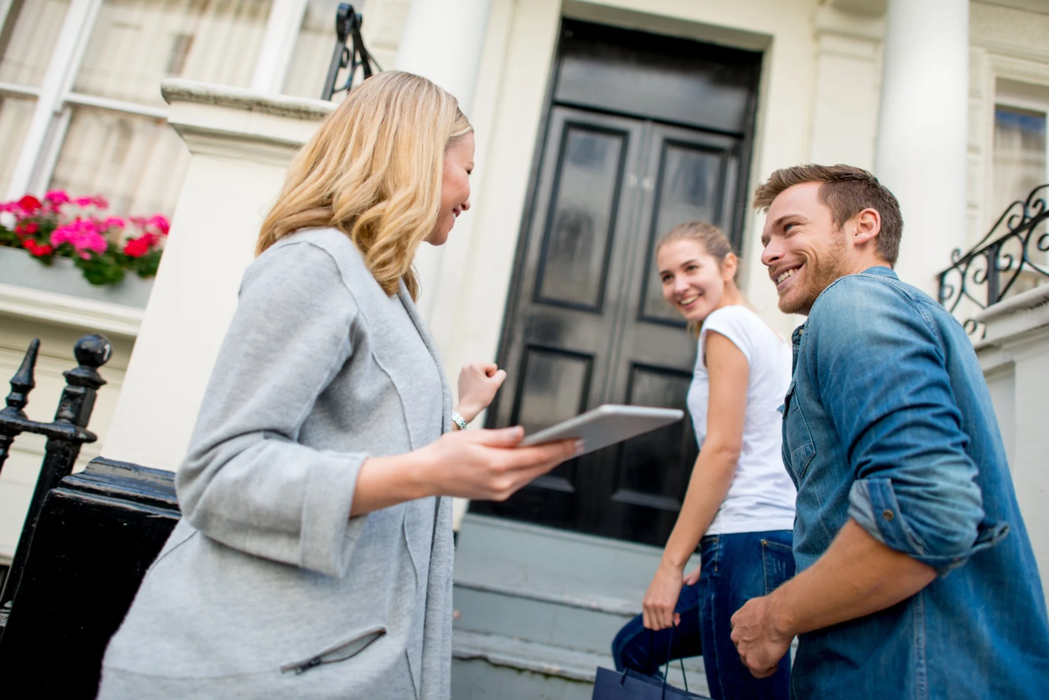 A woman is talking to a man and a woman in front of a house entrance. The woman is holding a notepad, and the man and woman are smiling. The house has a black door and flower boxes.