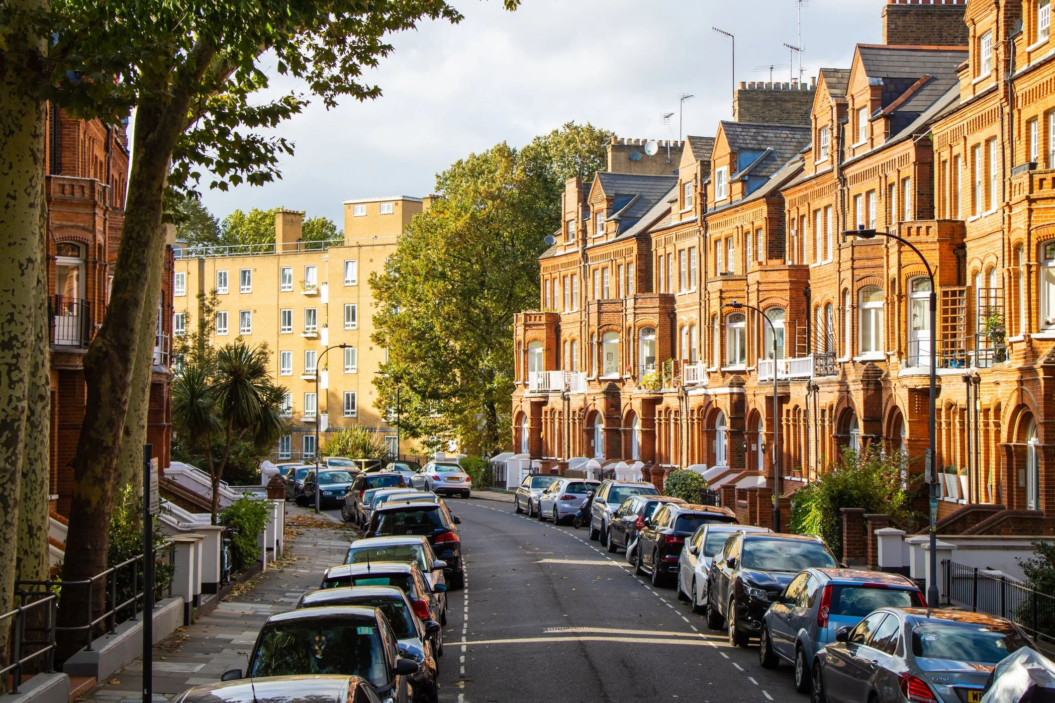 A street view with parked cars along a row of brick residential buildings with bay windows and balconies, tree-lined sidewalk, and a backdrop of modern apartment buildings under a cloudy sky.