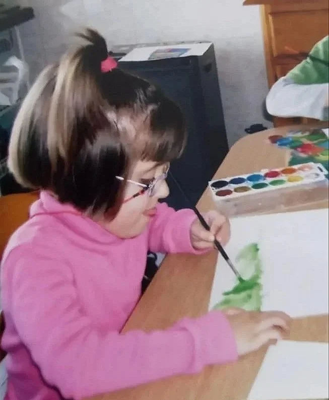 Young girl in pink sweatshirt painting with watercolor at table with watercolor set.