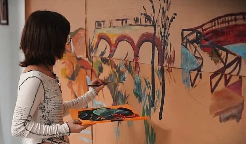 A woman with shoulder-length dark hair and glasses working on a large colorful mural of a bridge, trees, and landscape on a wall, holding a paint palette and brush.