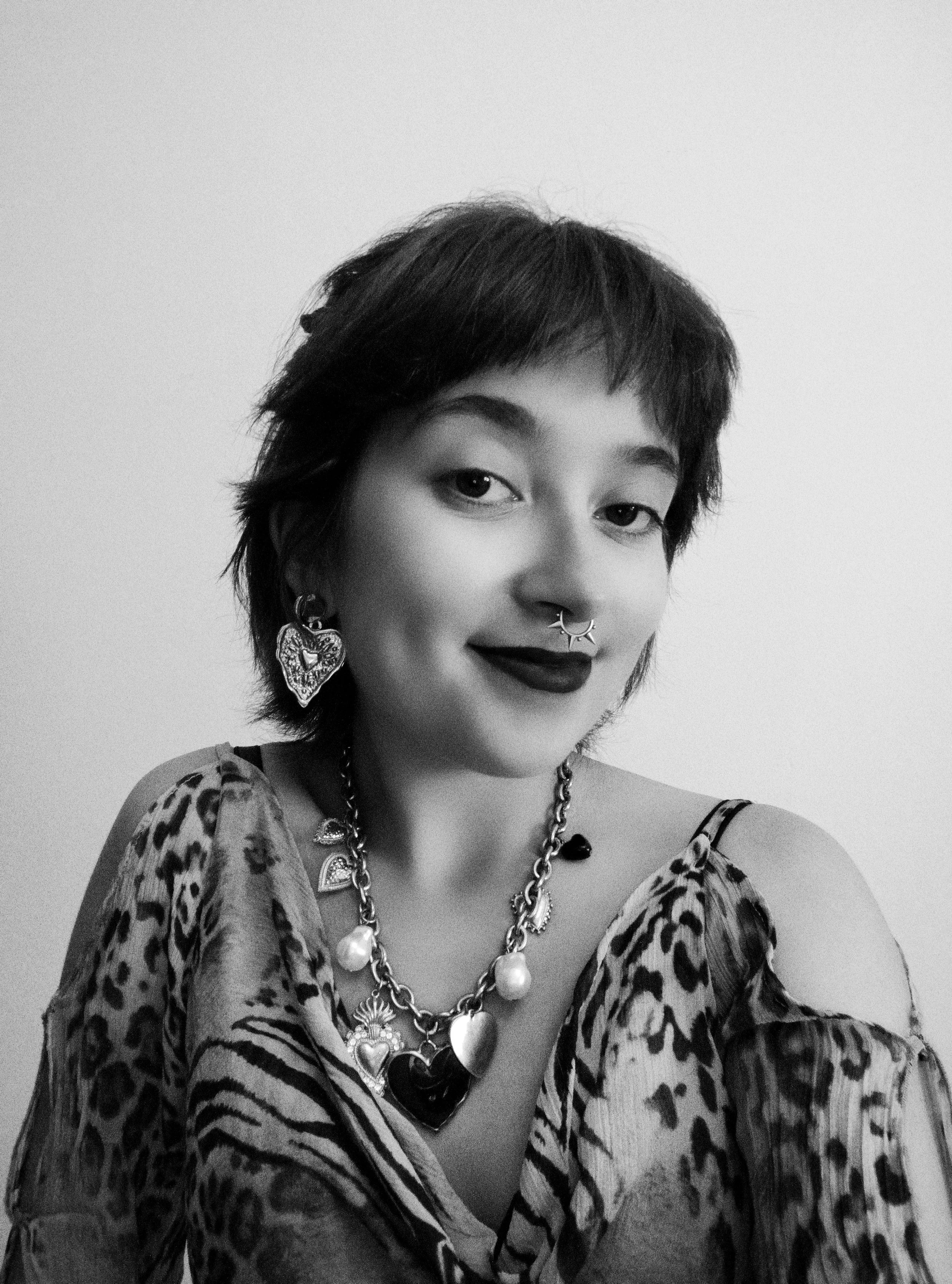 Black and white portrait of a young woman with short, tousled hair, wearing bold makeup, a leopard-print top with cold shoulders, and statement jewelry including large earrings, a layered necklace, and facial piercings.