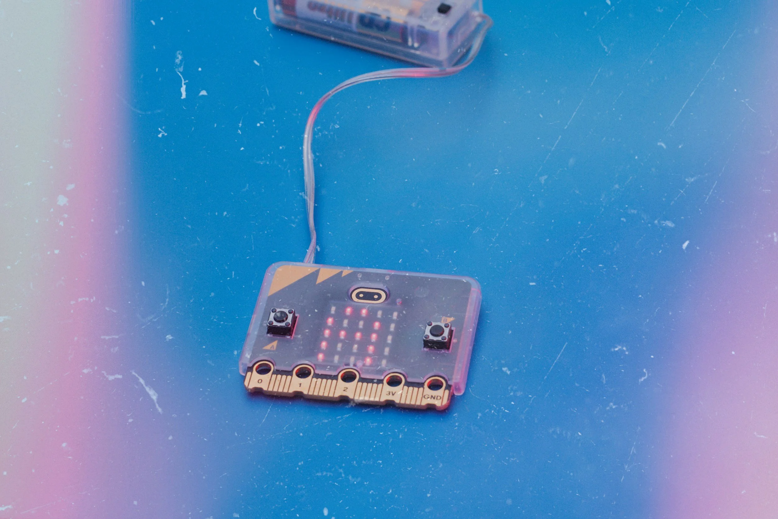 A small electronic circuit board with two black knobs, LED lights, and a series of labeled connection points, connected to a strip of tape and a larger clear plastic device, placed on a blue surface with visible scratches and a slight rainbow glow.