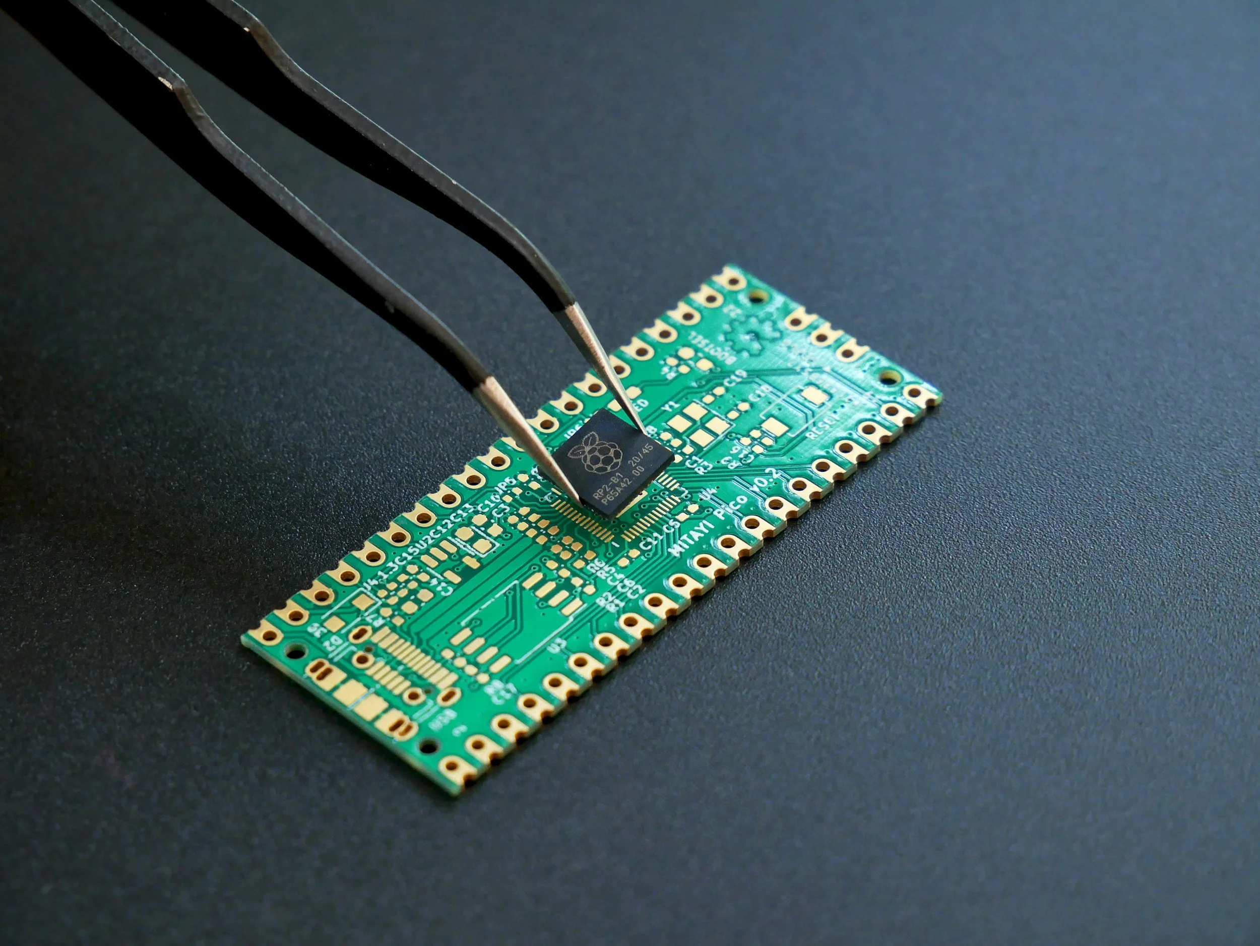 A pair of tweezers holding a black Raspberry Pi microcontroller on a green circuit board placed on a black surface.