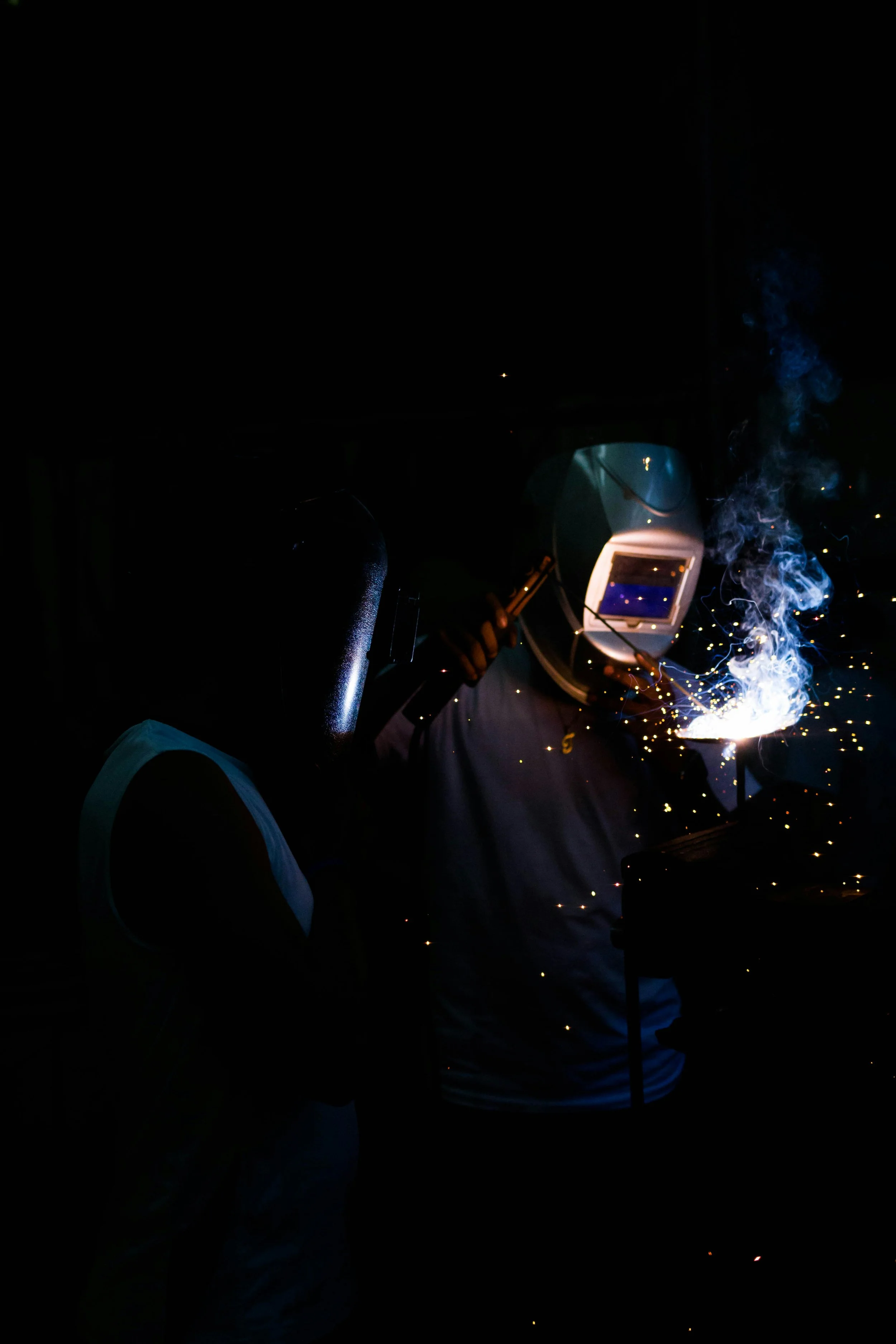 Two welders wearing protective gear working with welding equipment, sparks flying and smoke rising, in a dark environment.