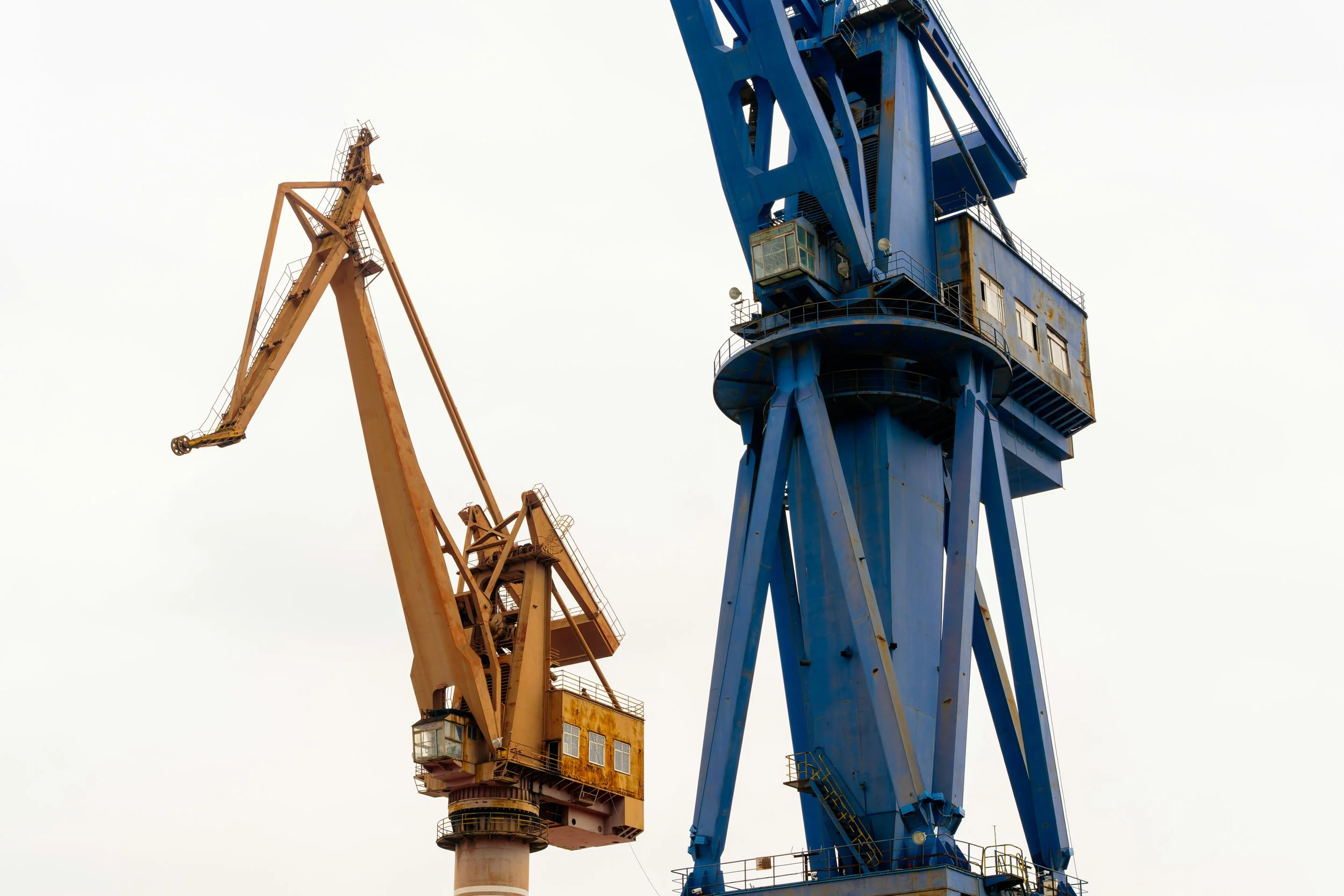 Close-up of two industrial cranes, one orange and one blue, against a cloudy sky.
