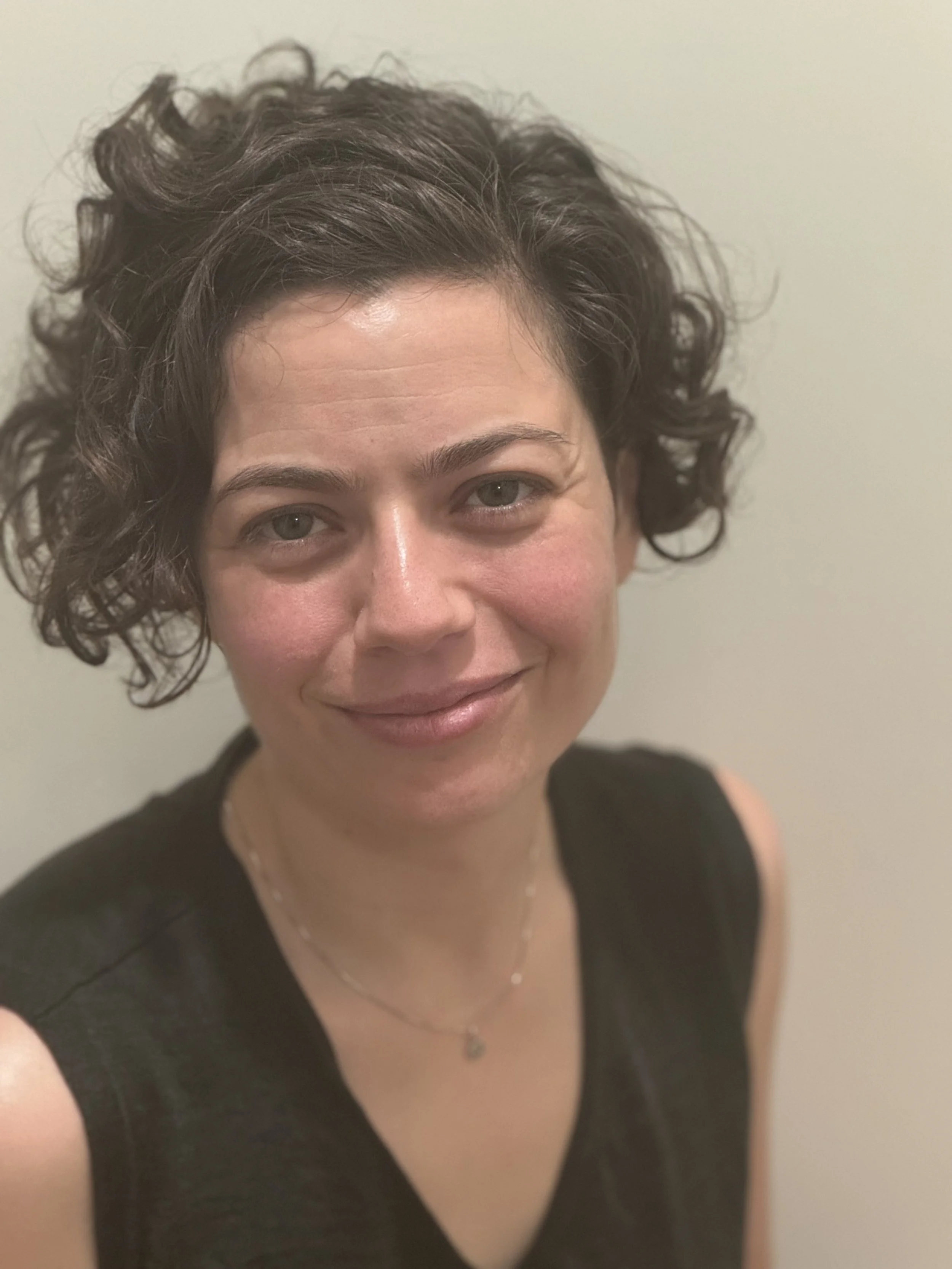 A woman with short, curly dark hair smiling at the camera, wearing a black sleeveless top and a delicate necklace.