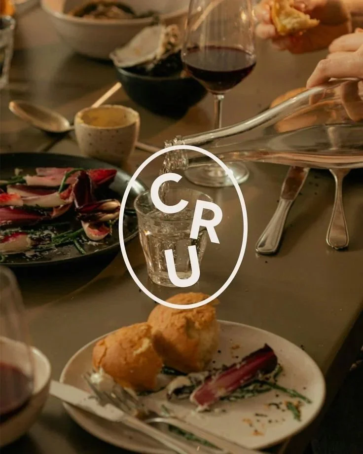 A dinner table with various dishes, including glass of red wine, fried food, and a plate with salad. A hand is pouring water into a glass with a logo that reads 'CRU' over it.
