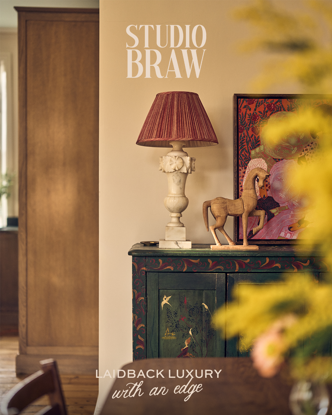 Decorative interior scene with a dark green painted cabinet adorned with bird and plant motifs, topped with a vintage table lamp with a red pleated shade, a carved wooden horse sculpture, and a colorful framed artwork in the background, suggesting a cozy, elegant living space.