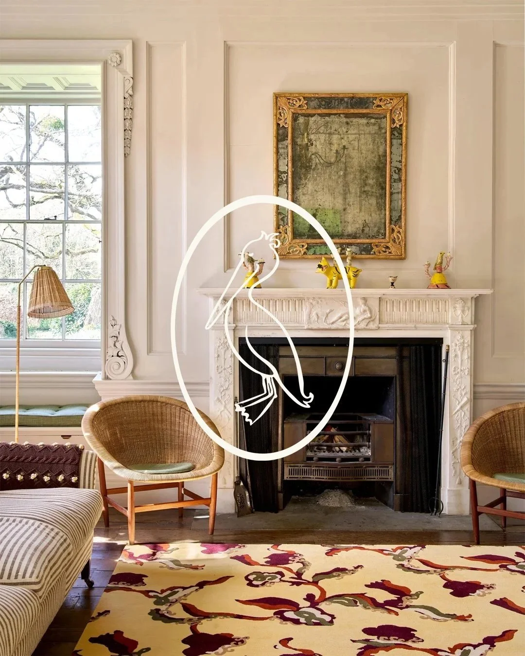 Living room with white ornate fireplace, large window on the left, and two rattan chairs. A colorful floral rug is on the wooden floor, and a large framed painting hangs above the fireplace. Decorative figurines on the fireplace mantel include yellow and pink figures, and a folding fire grate is in the fireplace.