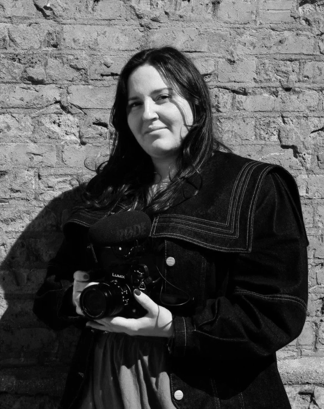 Young woman with dark hair holding a camera and a microphone, standing against a brick wall.