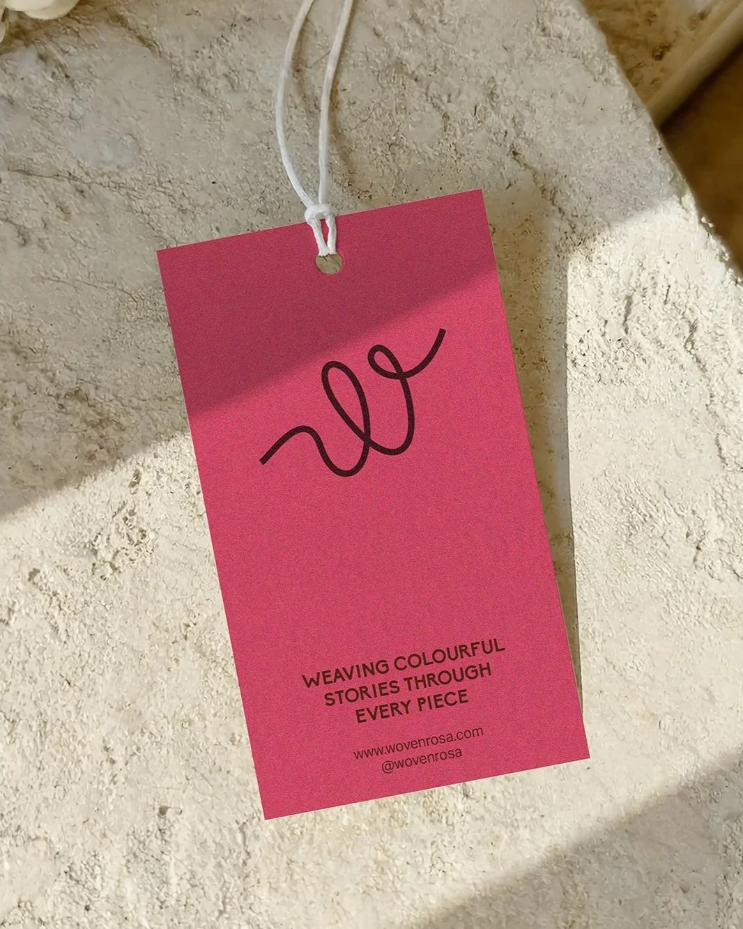 A pink tag hanging on a textured beige wall with a black cursive logo and the text 'Weaving colourful stories through every piece' along with a website and social media handle.
