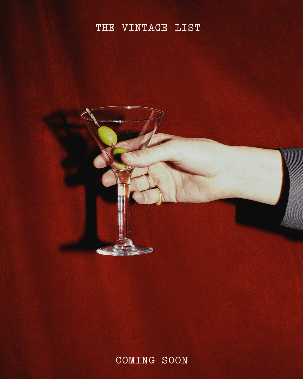 A hand holding a martini glass with green olives against a red background, with the text 'The Vintage List' at the top and 'Coming Soon' at the bottom.
