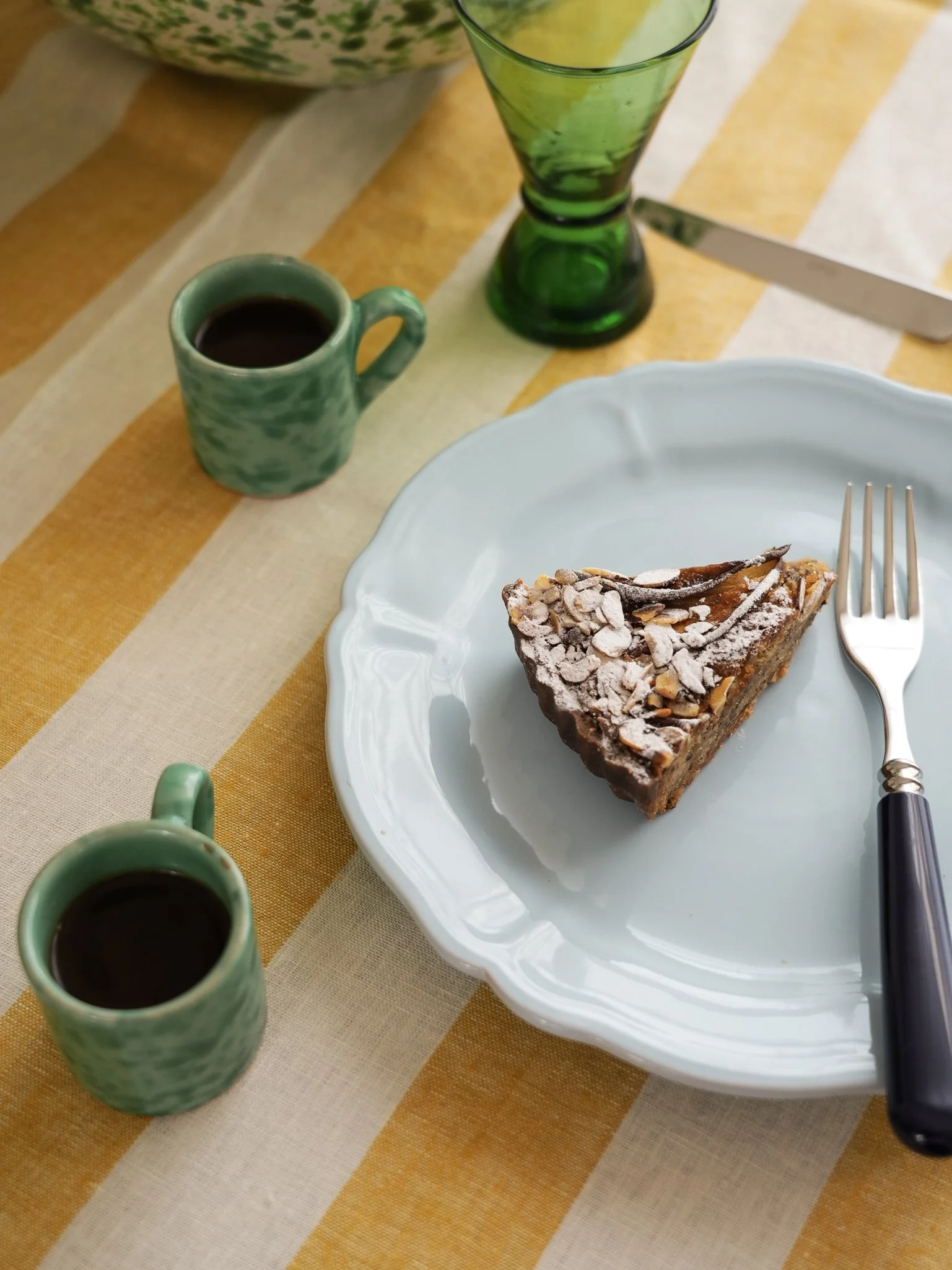 A piece of chocolate cake with nuts and powdered sugar on top on a white decorative plate, accompanied by a fork. Two green coffee mugs filled with black coffee, a green glass of water, and a green ceramic pitcher are on a yellow and white striped tablecloth.