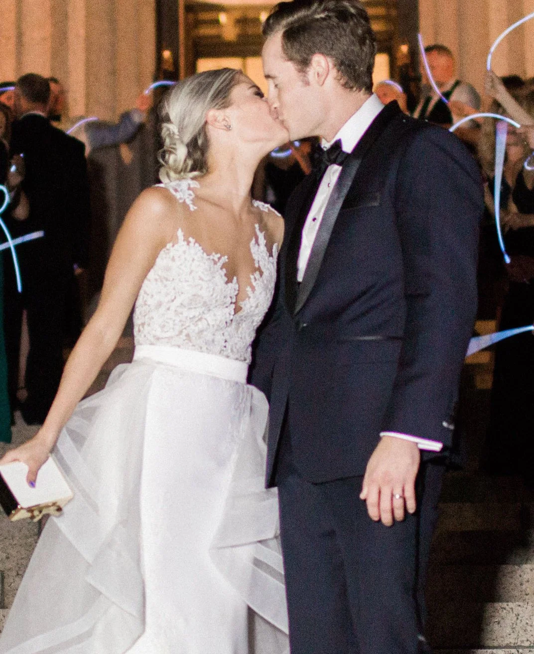 A bride and groom kiss at their wedding reception, dressed in formal attire. The bride is wearing a white lace wedding gown with a tulle skirt, and the groom is in a black tuxedo with a bow tie. They are surrounded by guests at the celebration.
