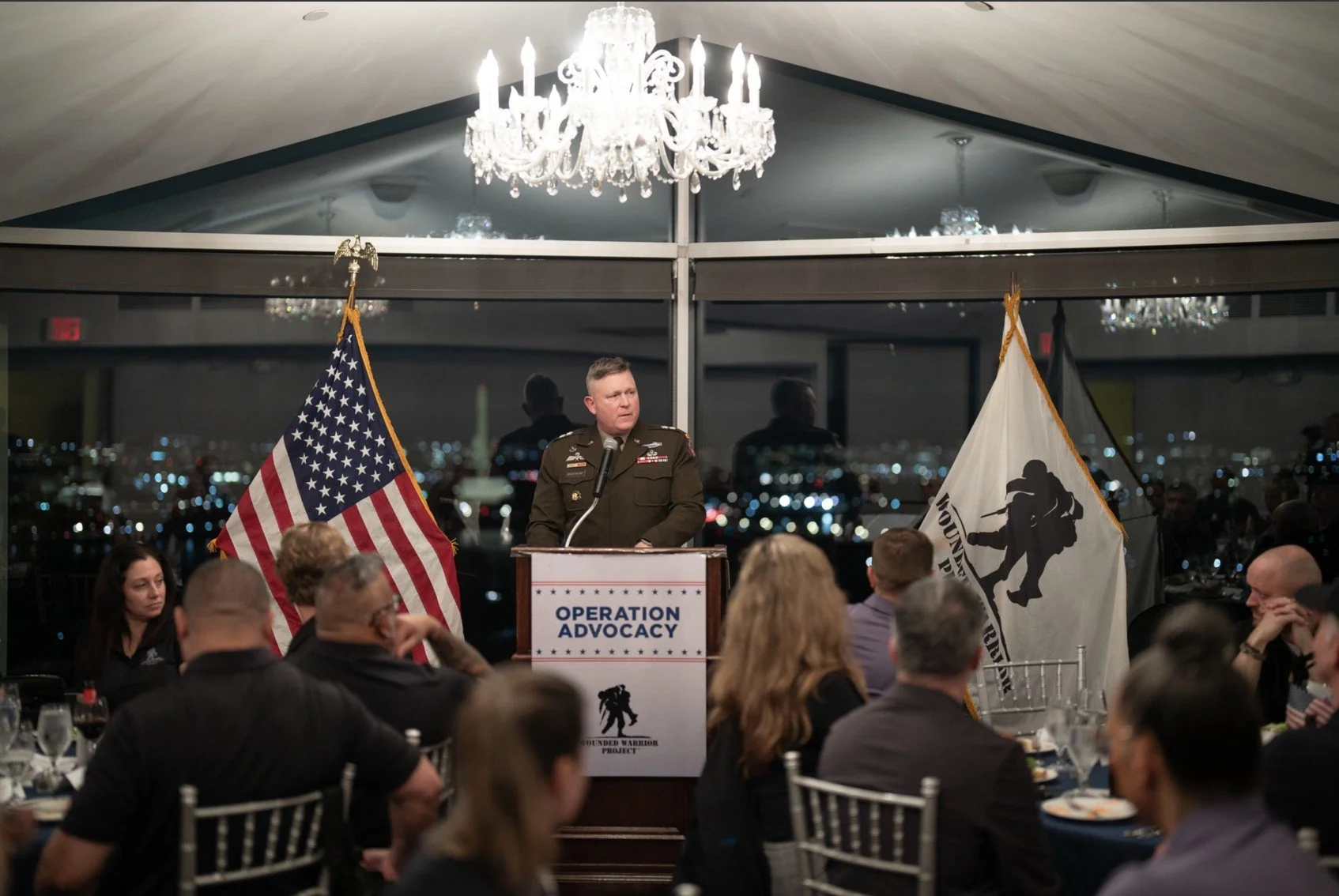 A military officer speaking at a podium during an event called Operation Advocacy, with the American flag and a second flag behind him, in a room with a view of city lights at night.