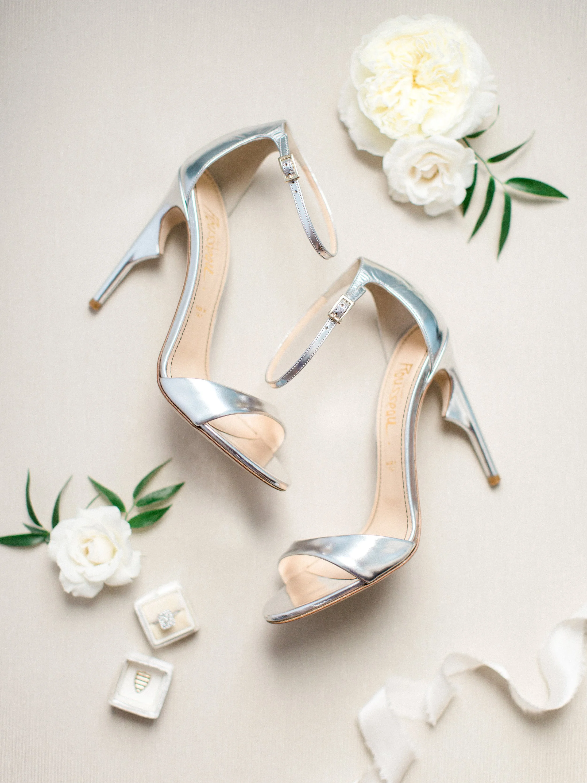 A pair of silver high-heeled shoes with ankle straps, white flowers, a mounted ring, and a small box on a light surface.