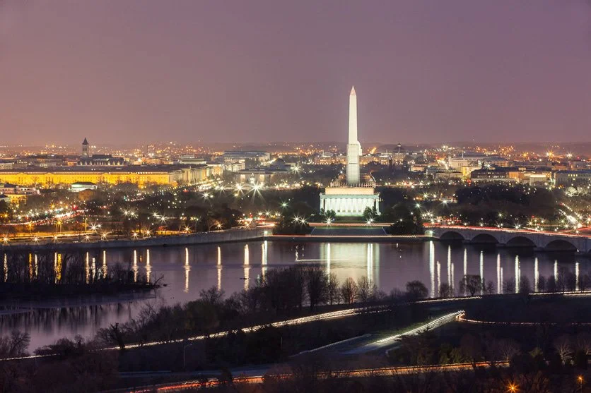 Nighttime view of Washington D.C. featuring the Washington Monument illuminated next to the reflecting Lincoln Memorial Reflecting Pool with city lights in the background.