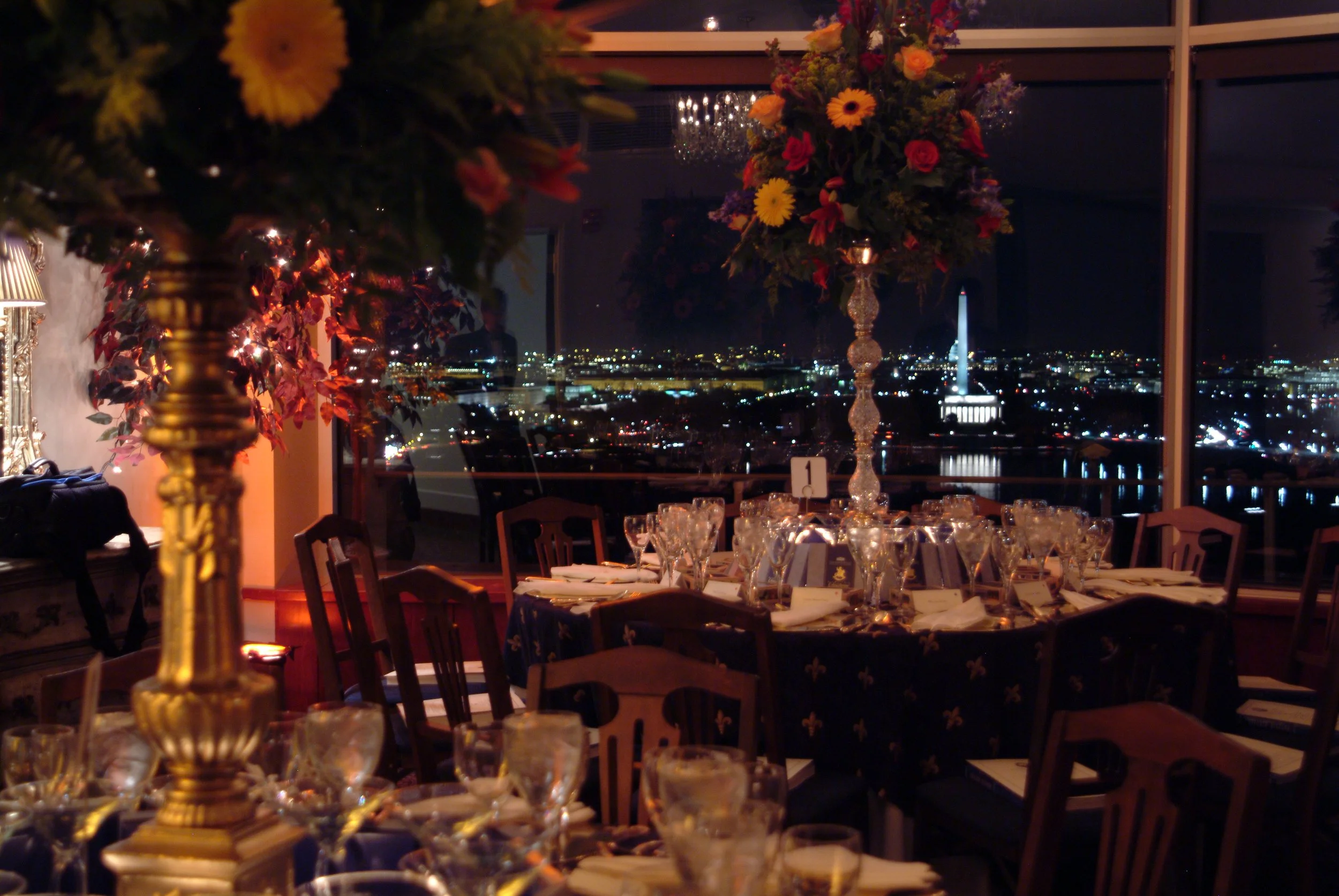 Elegant dinner setup with large floral centerpieces on tables, overlooking a city skyline at night through large windows.