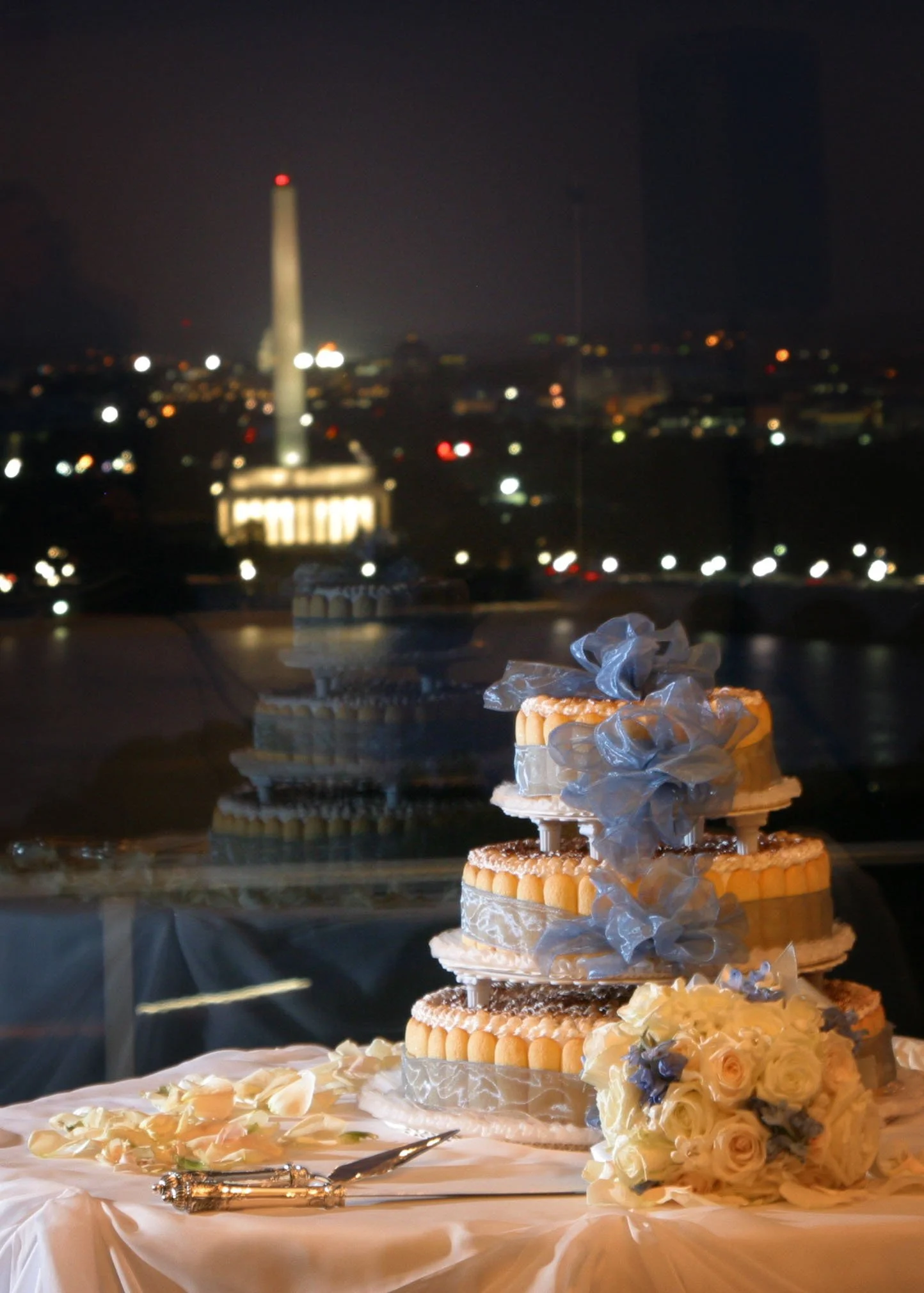 A multi-tiered wedding cake with floral and ribbon decorations, placed on a table with a bouquet of white and cream roses, against a window showing the illuminated Washington Monument and cityscape at night.