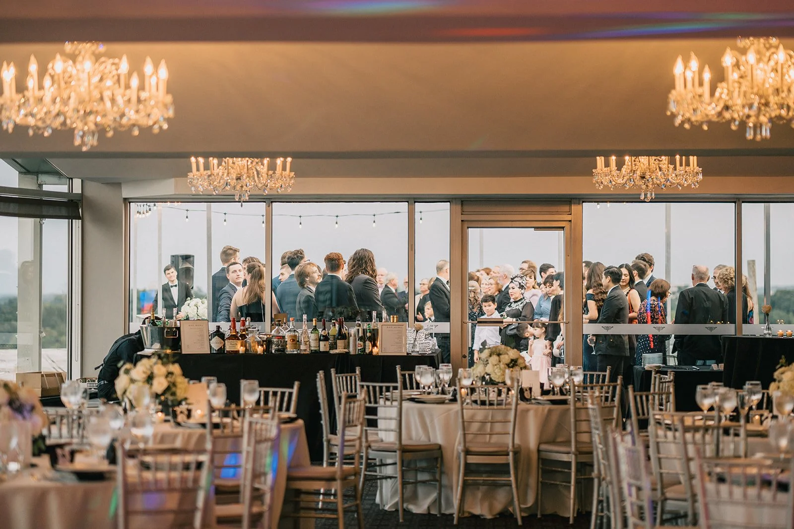 Guests at a wedding reception gather outside on a balcony, seen through large glass windows, while the indoor area is decorated with chandeliers, round tables with beige tablecloths, and floral centerpieces.