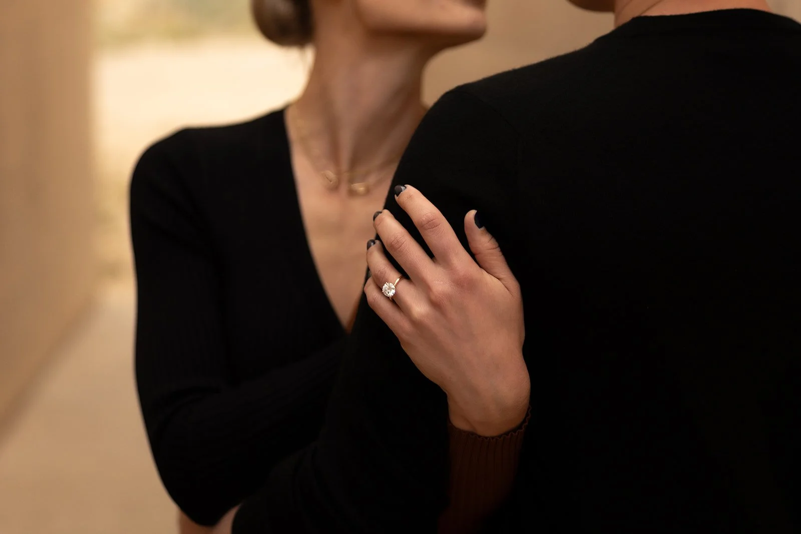 Close-up of a woman with a ring on her finger touching the shoulder of a man, both dressed in black, indoors with soft lighting.